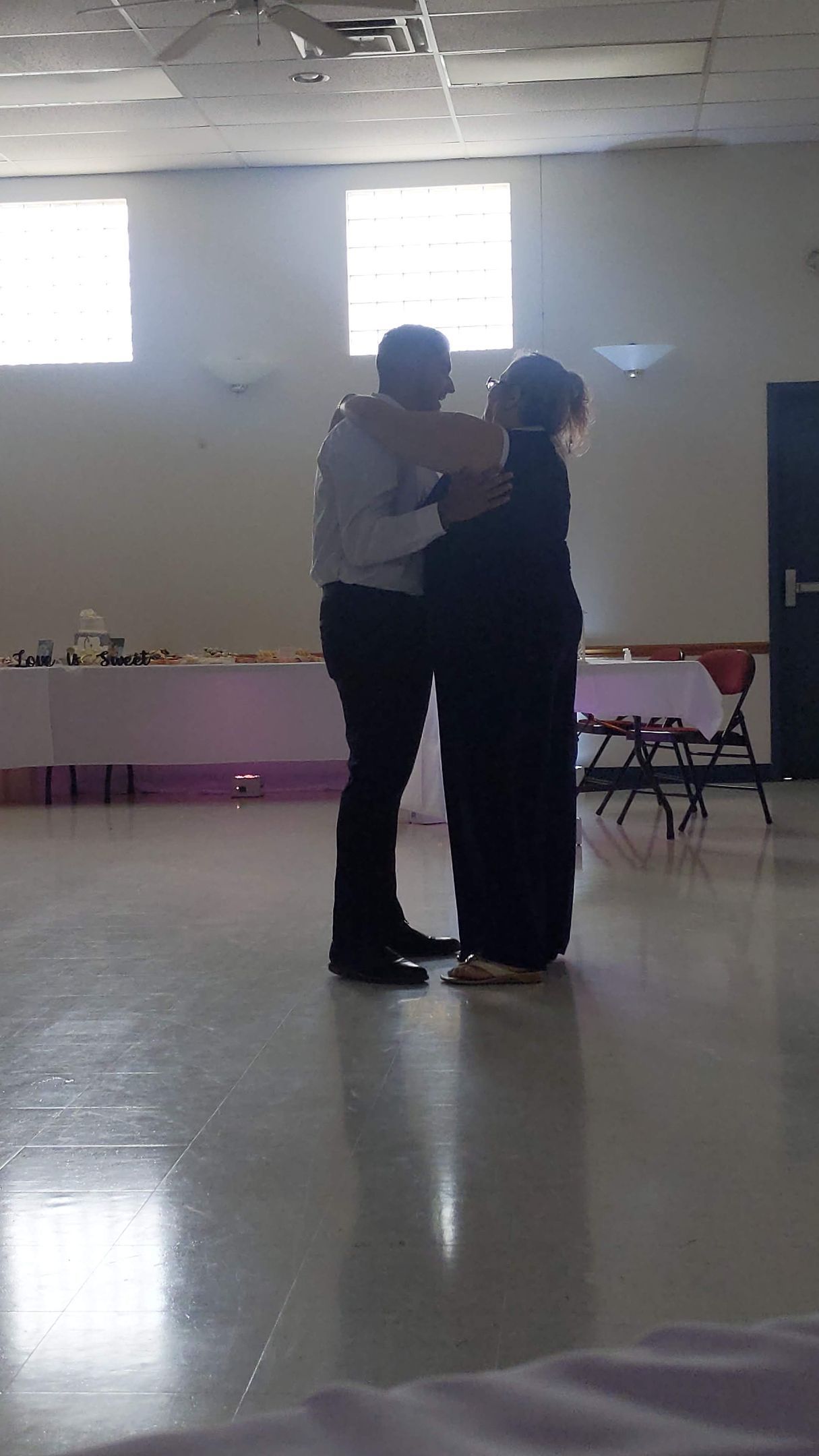 Two people dancing together in a bright hall with polished floors and tables along the back wall.