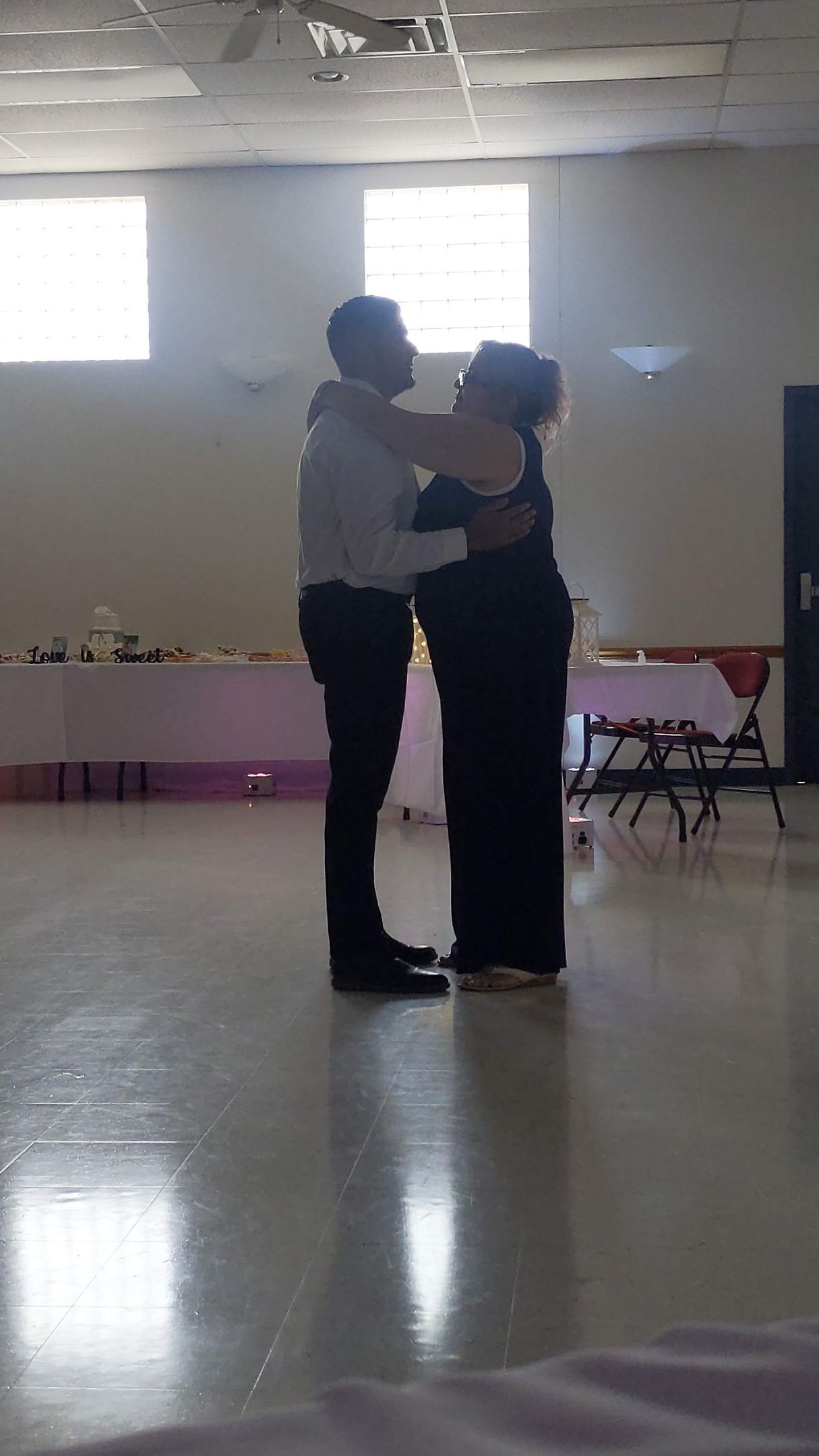 Two people embracing on a shiny floor in a bright hall with tables along the wall.