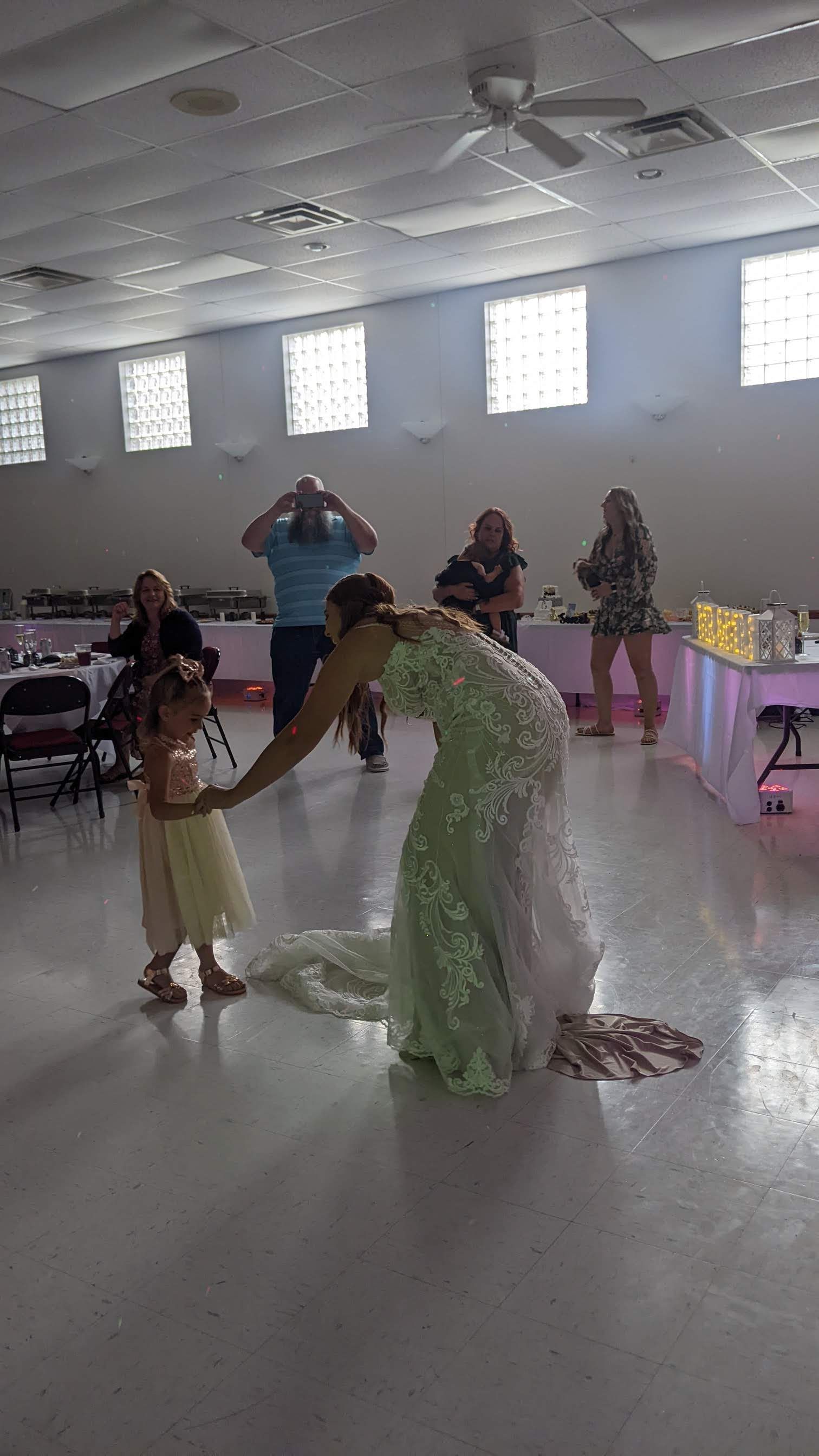 Costumed performer in a green dress bending to a child in a yellow dress at a bright indoor event.