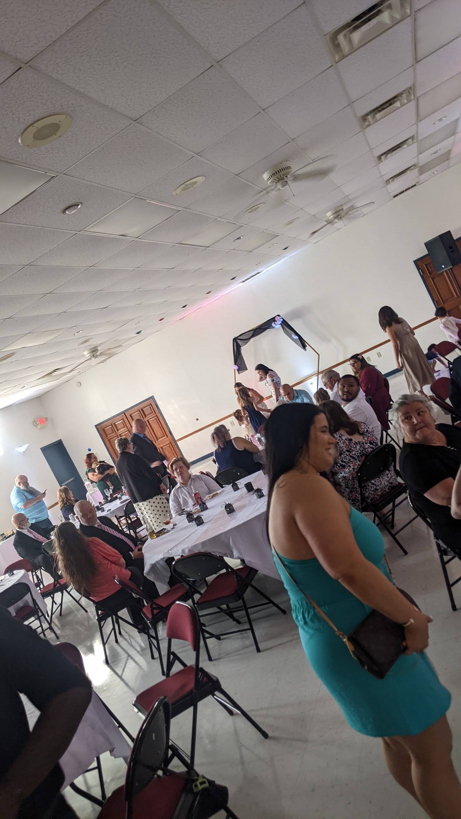 Indoor event hall with people seated at tables and a woman in a teal dress standing in the foreground