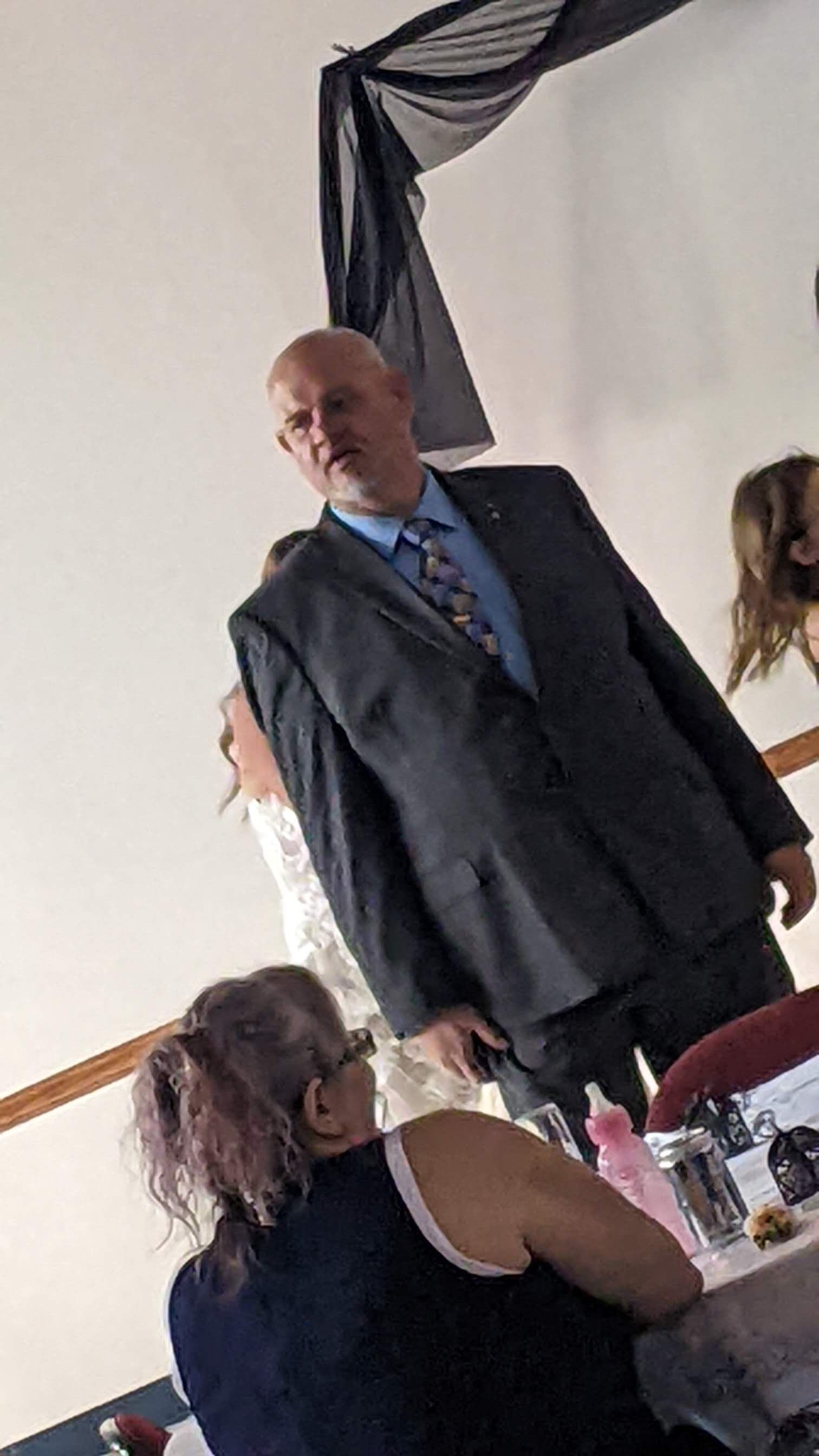 Man in a dark suit stands and speaks at a decorated indoor event, with seated guests nearby.