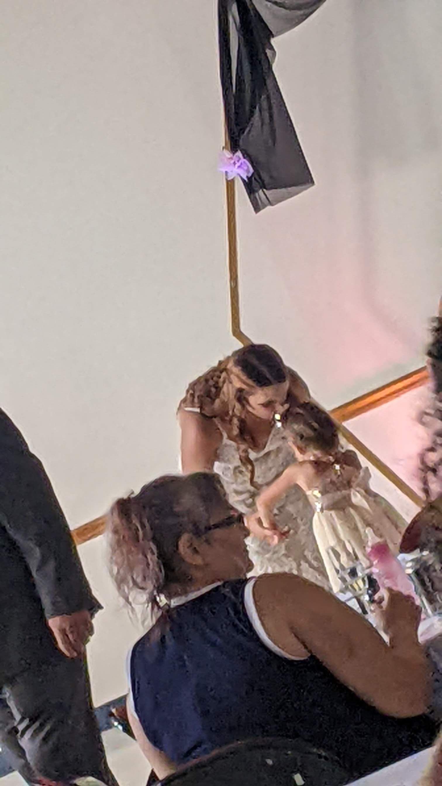 Woman in a cream dress crouches beside a seated child at an indoor event with guests nearby.