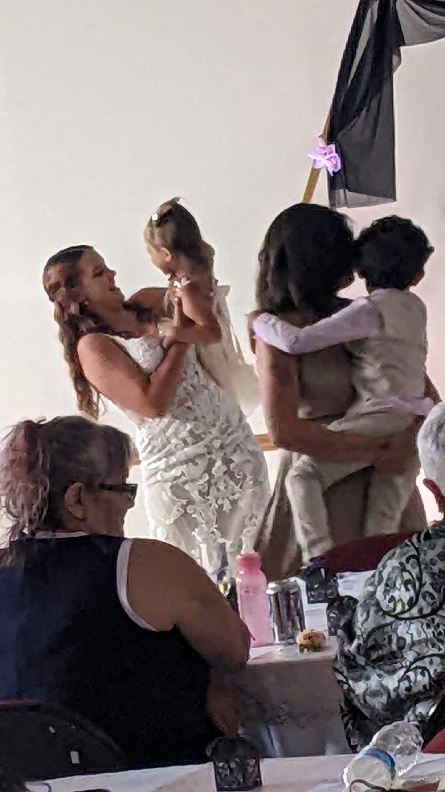 Two people embracing at a formal indoor event, with one in a white dress and guests seated nearby.