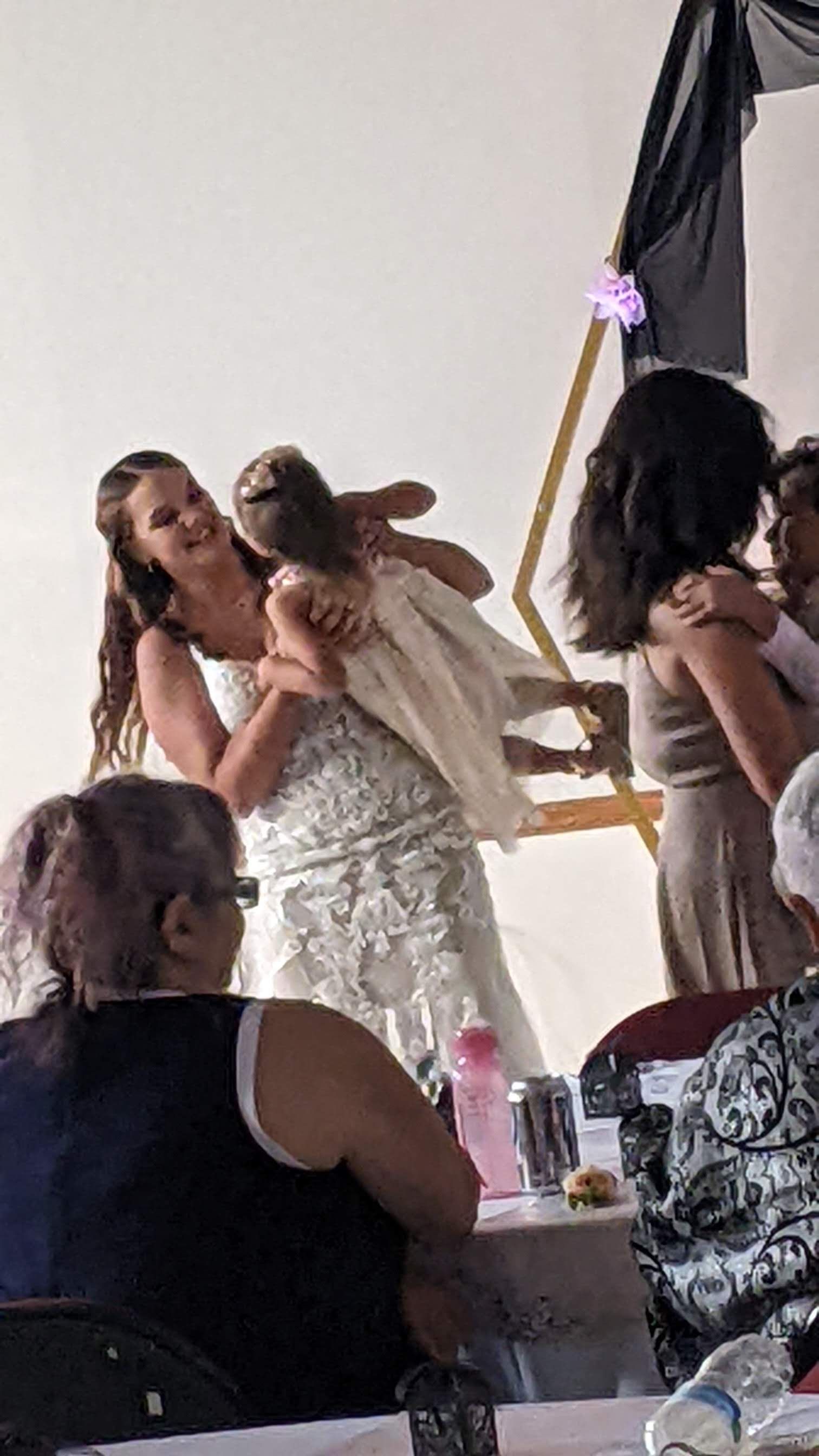 People posing in white outfits at an indoor celebration, with one holding a baby and a black drape in the background
