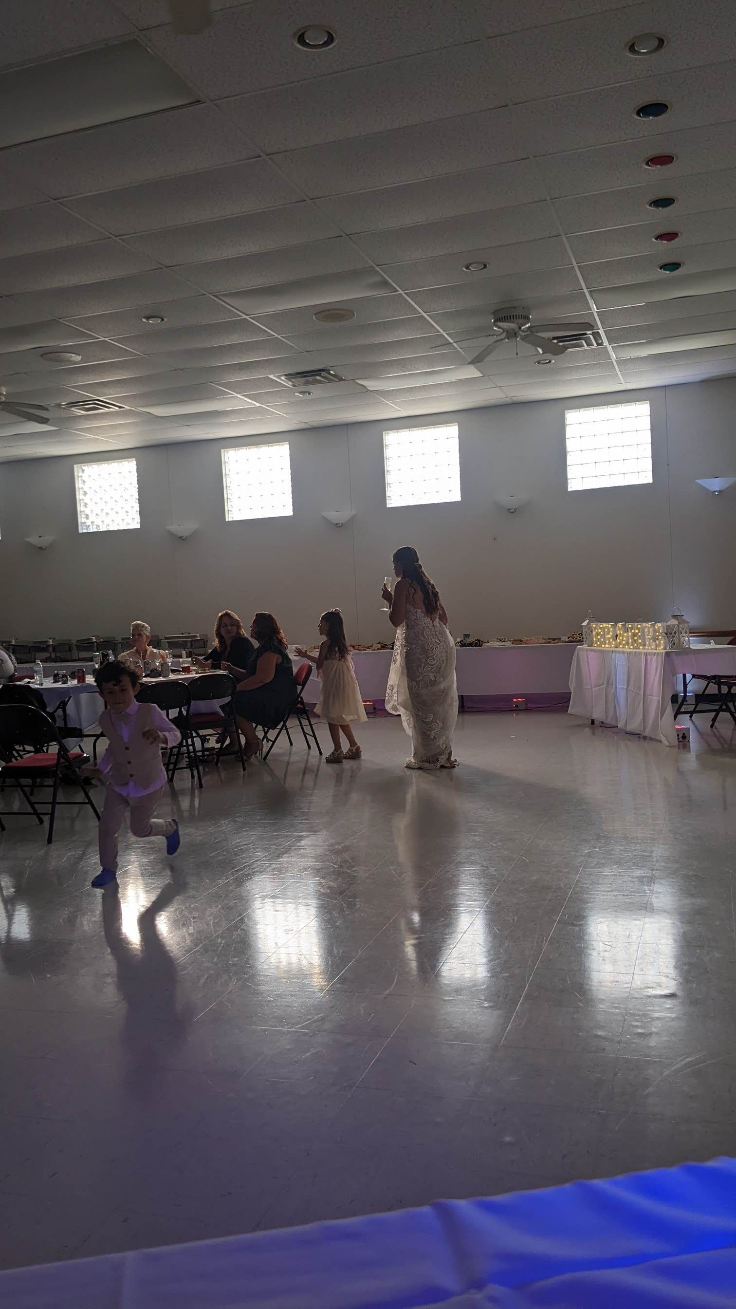Indoor banquet hall with a few people seated and standing near decorated tables, reflected on a shiny floor.