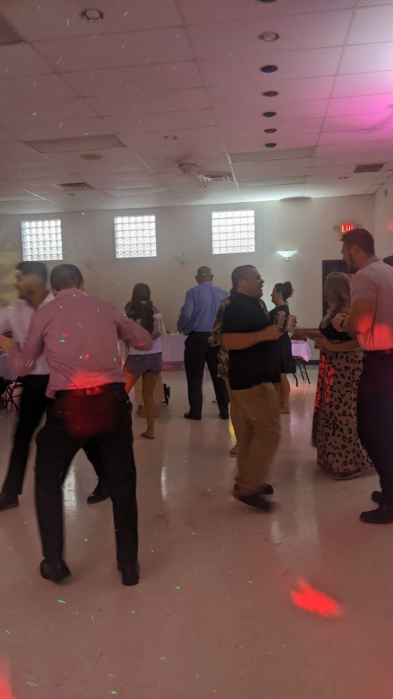 People dancing at a party in a bright hall with red and pink lights.