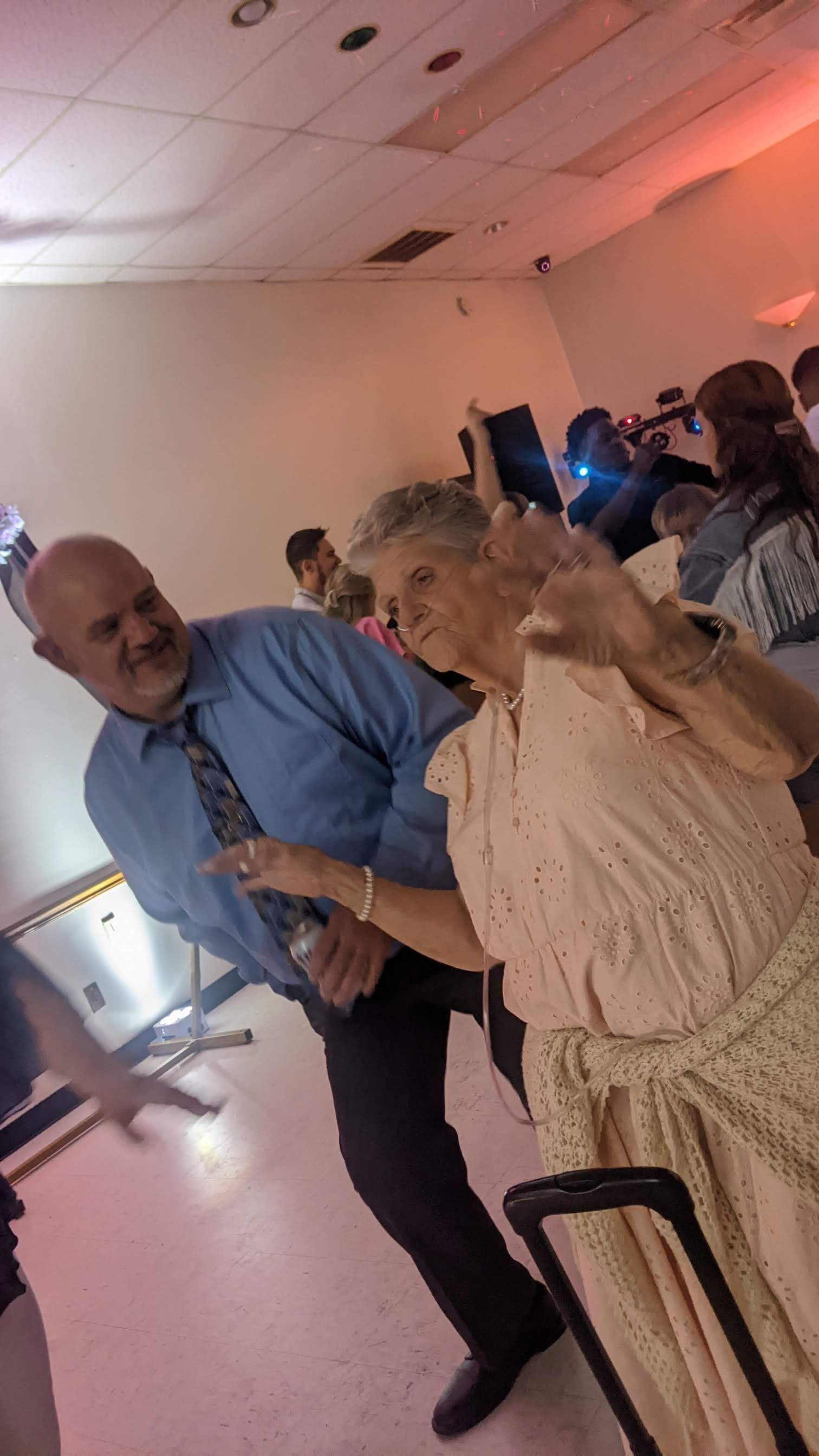 Two people dancing at a party, smiling under pink lighting in a crowded room.