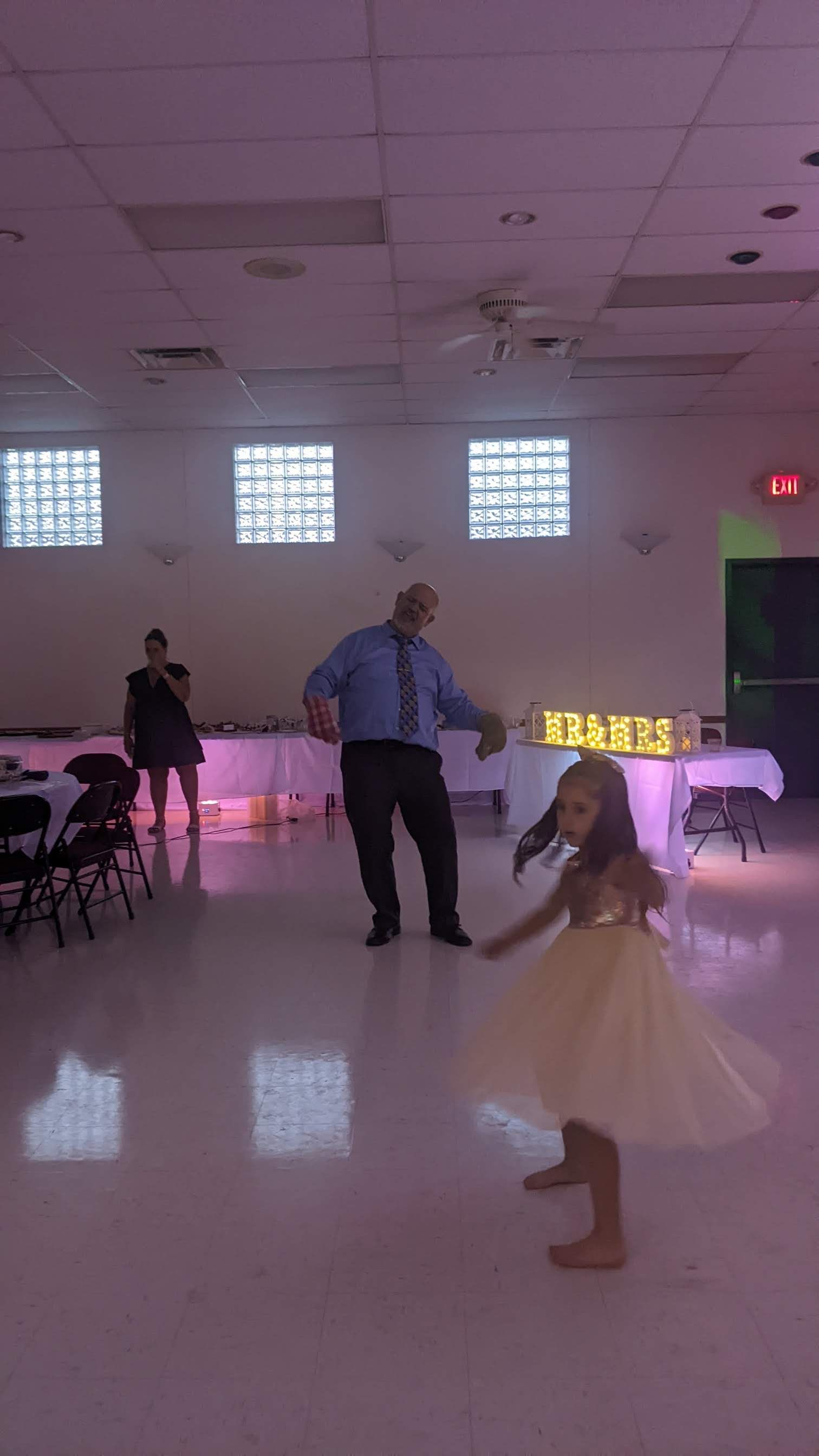 Dance floor with a child in a white dress, an adult, and pink-lit tables in a banquet hall