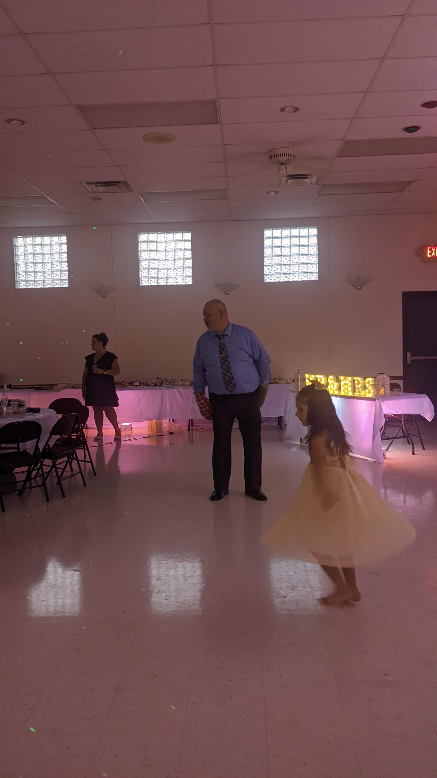 A man in a blue shirt stands on a pink-lit dance floor while a child in a yellow dress dances nearby.