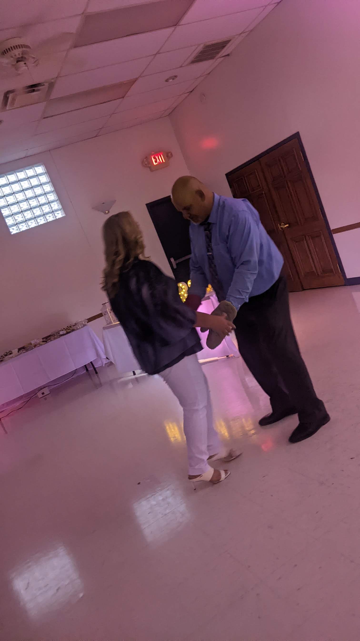 Two people dancing in a pink-lit room with a tiled floor and wooden door.