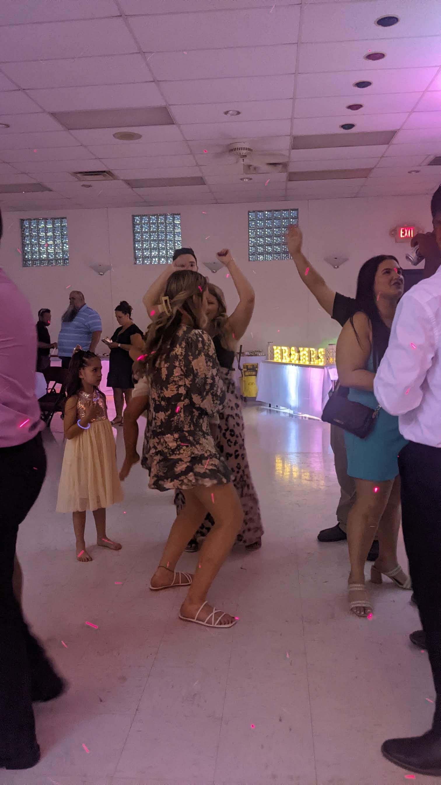 People dancing at a party in a bright hall, with guests mingling in the background.