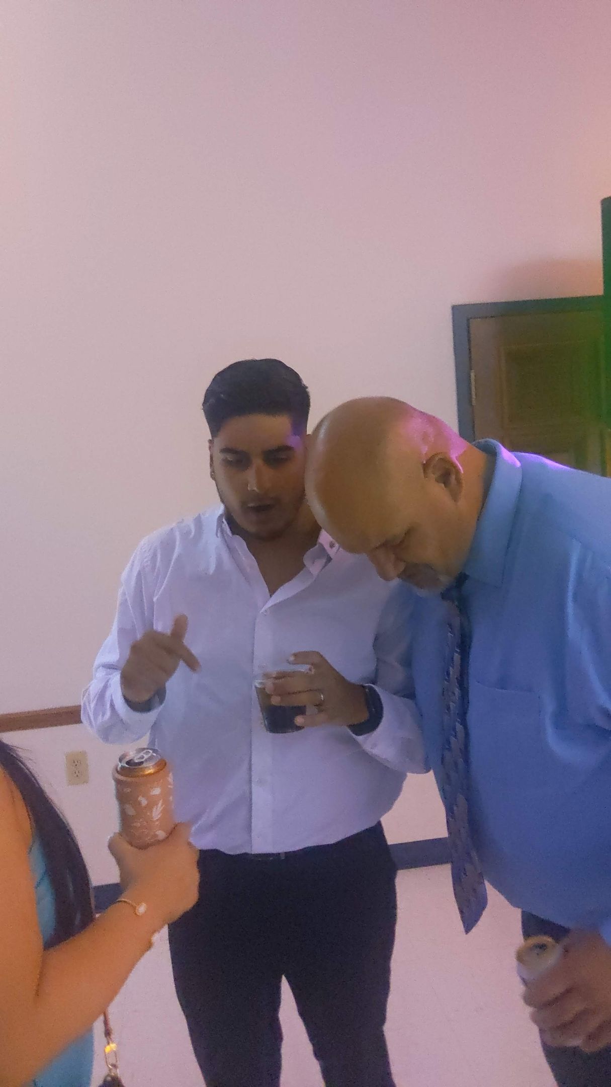Two men indoors, one in a white shirt gesturing while the other in a blue shirt looks down at a phone.