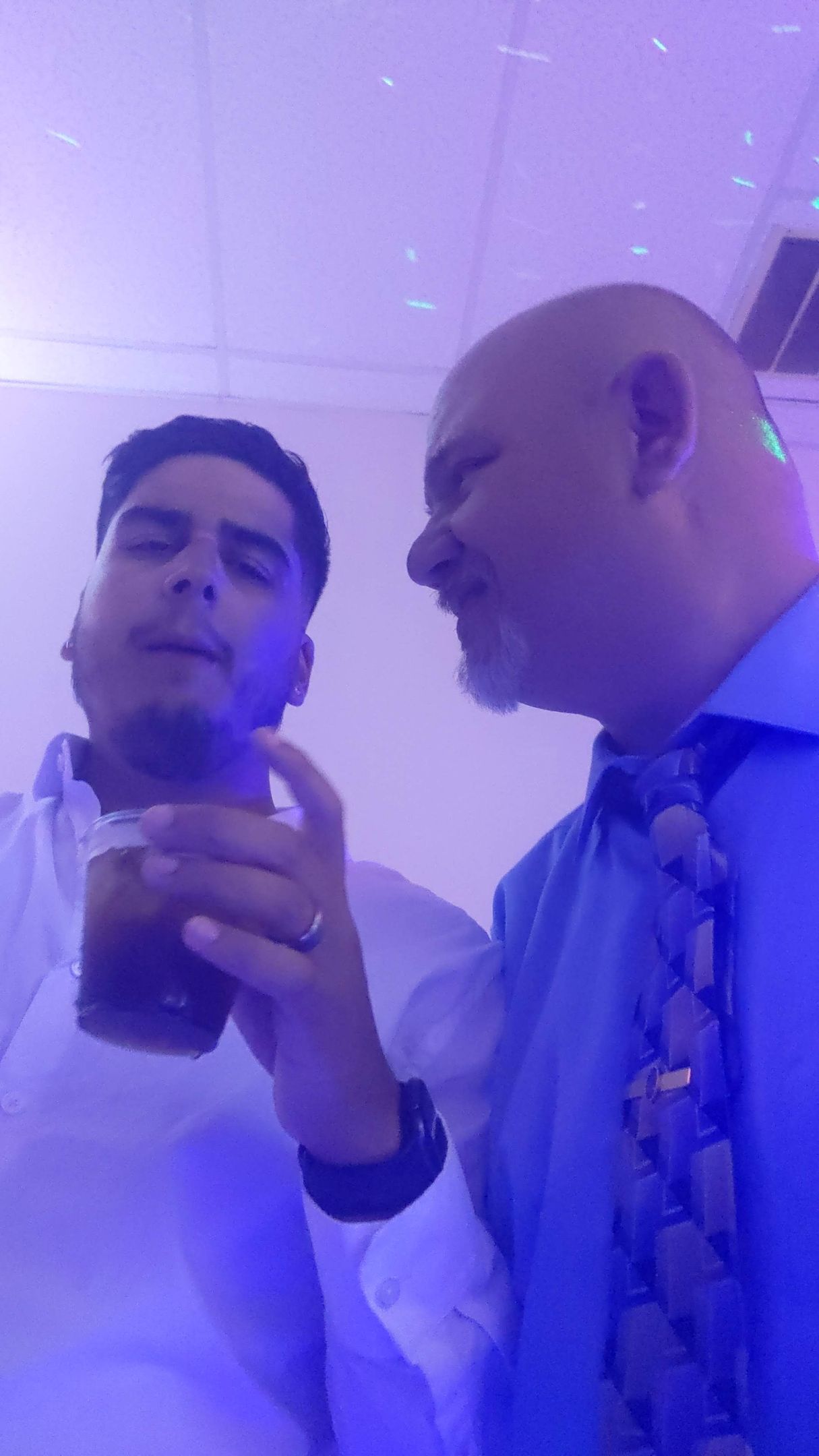 Two people in dress shirts clink drinks under purple party lighting.