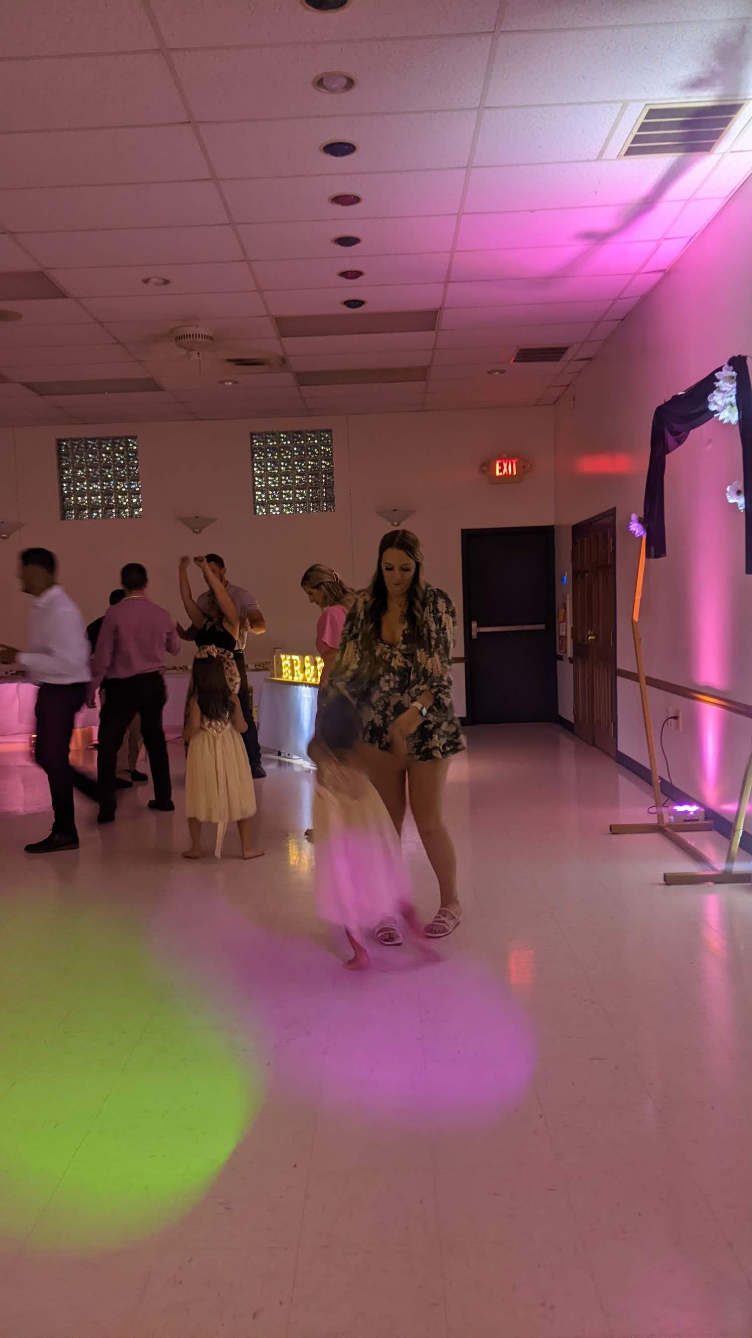 People dancing in a dimly lit hall with pink and purple lights, a child in the foreground.