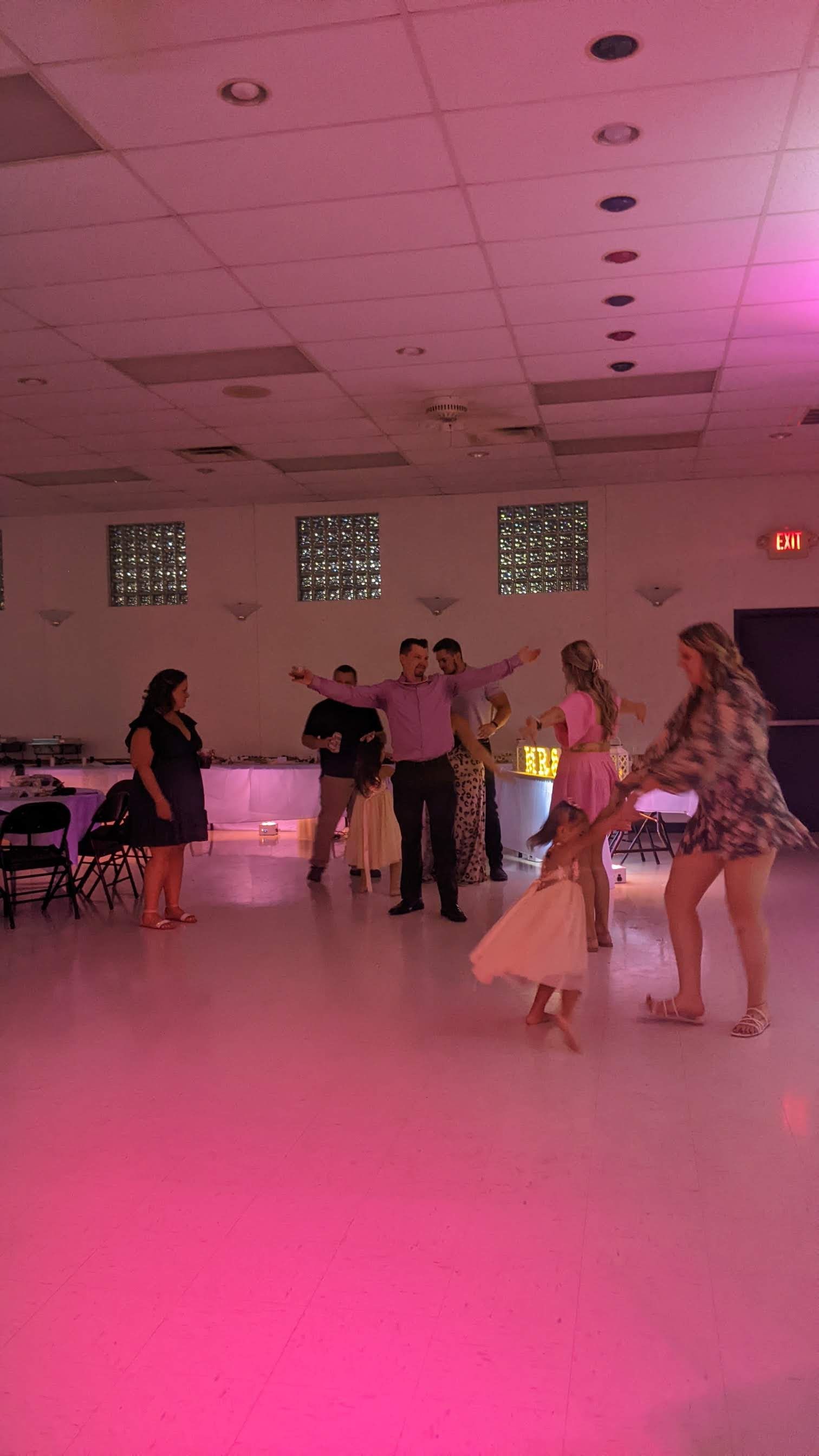 People dancing in a pink-lit hall, with tables and chairs along the sides.