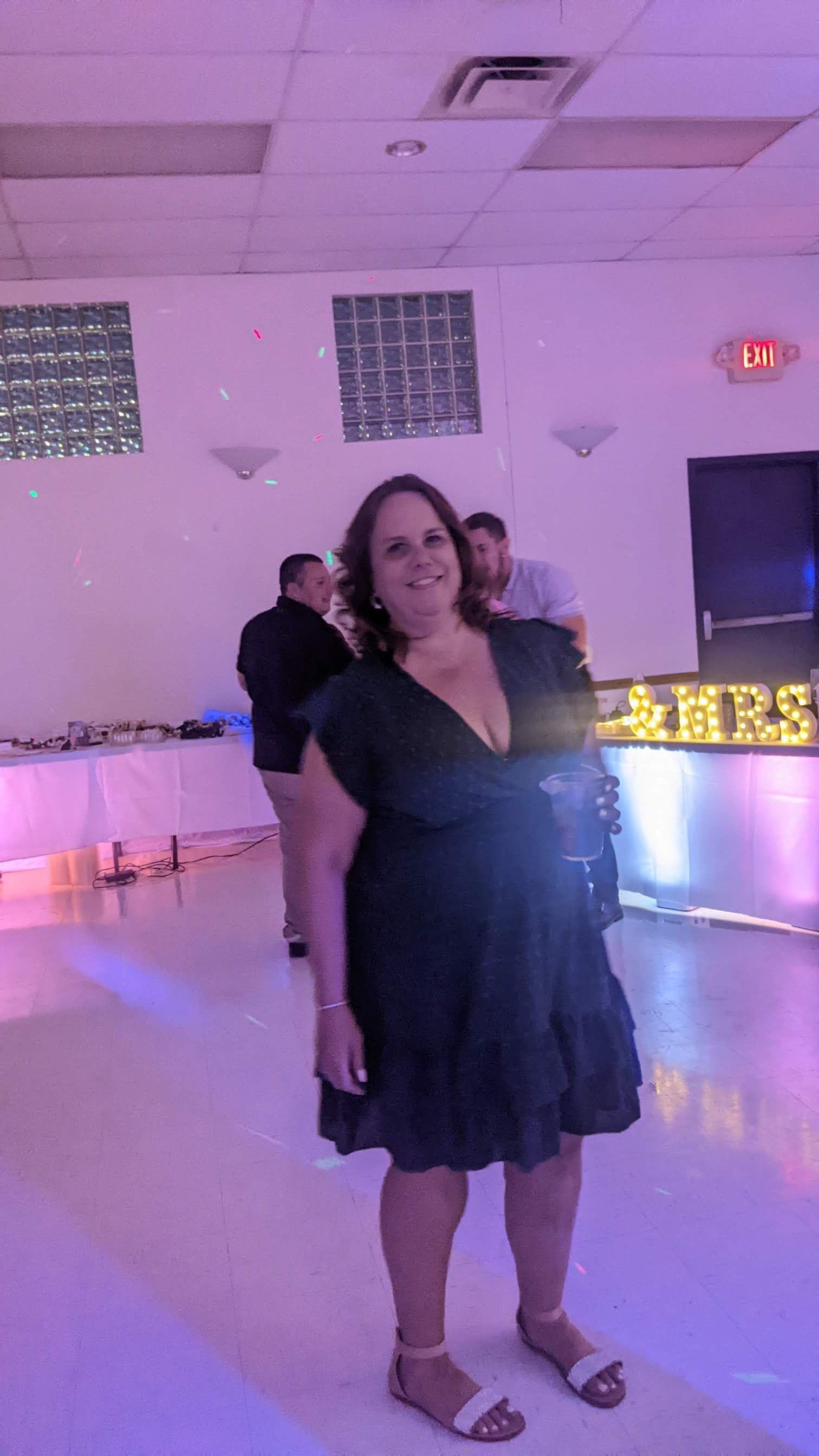 Woman in a black dress standing in a lit party venue with glowing letters and a dance floor behind her