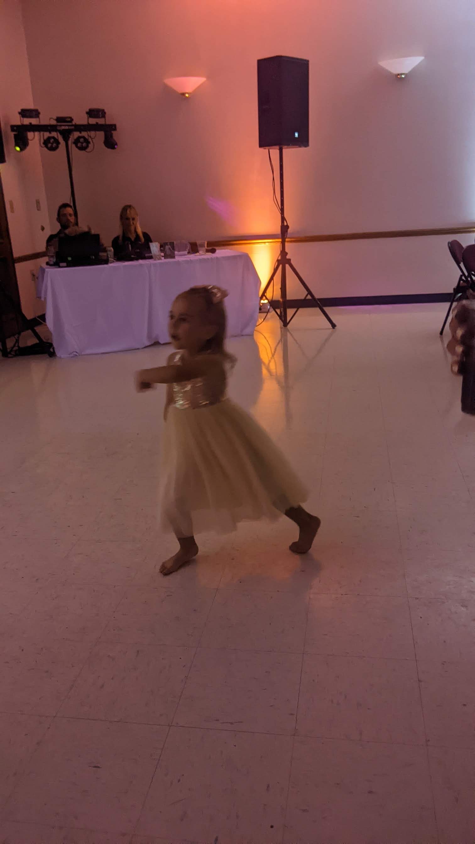 Child in a cream dress dancing on a lit indoor floor during a celebration