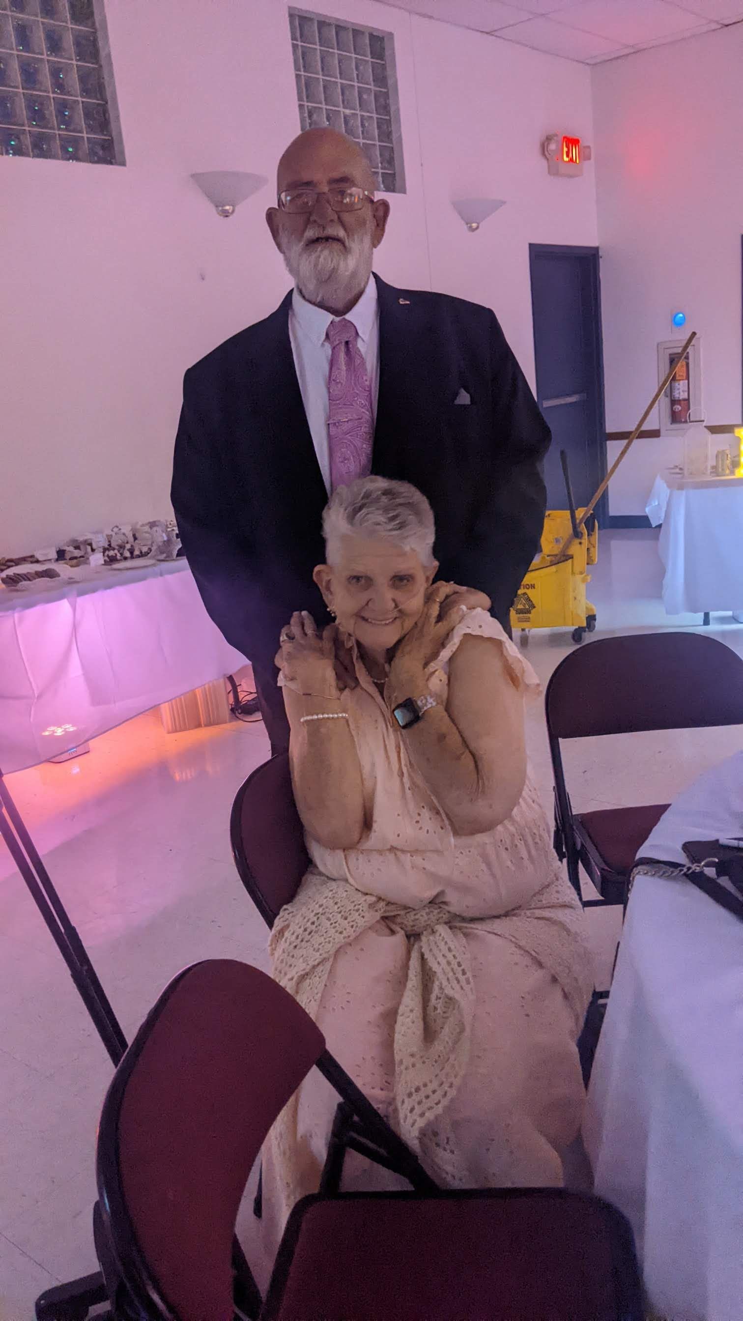 Two people posing at a formal event, with one standing behind a seated guest in a decorated indoor venue