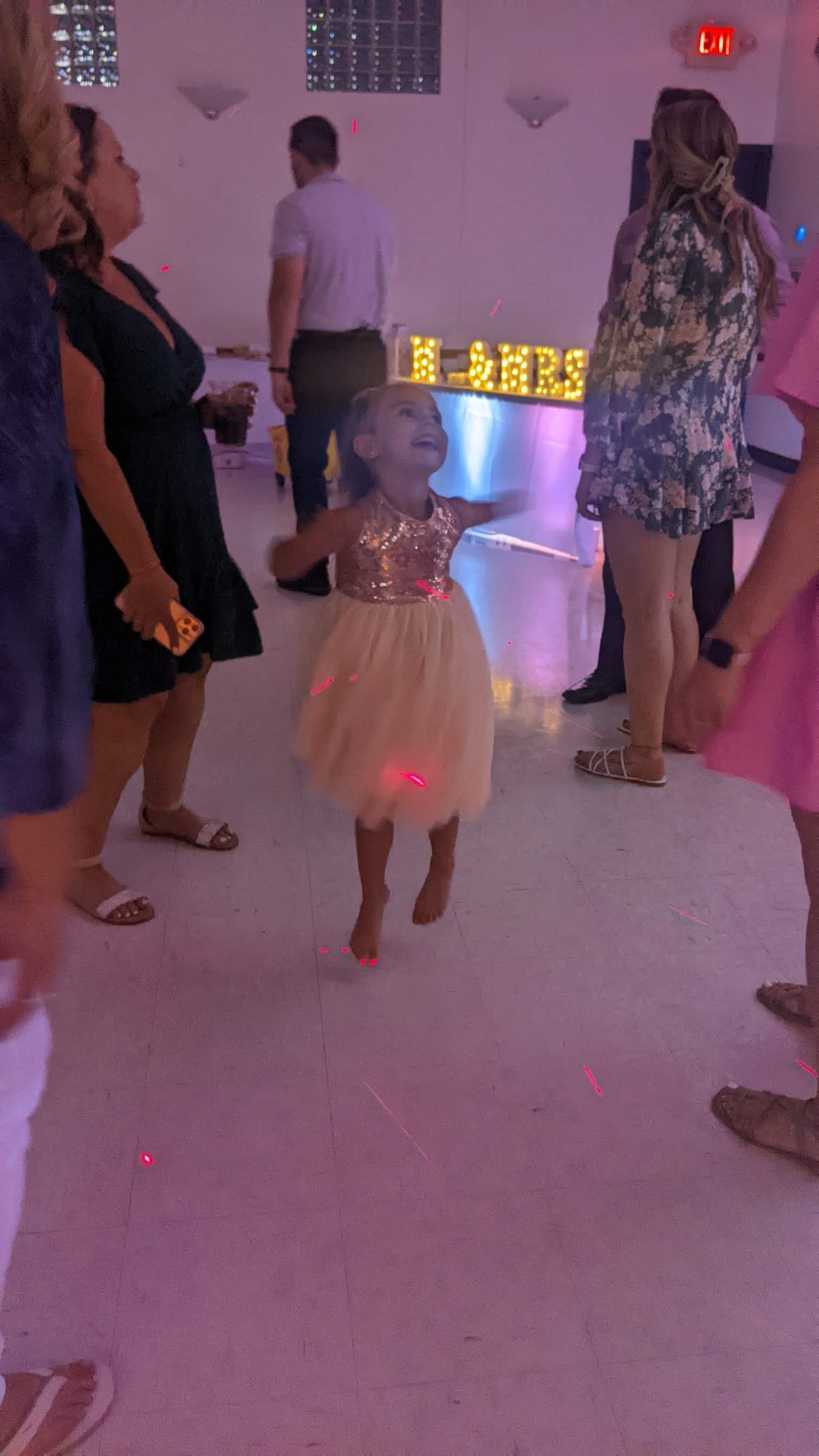 Child in a white dress dancing at a party, surrounded by adults in a softly lit indoor space.