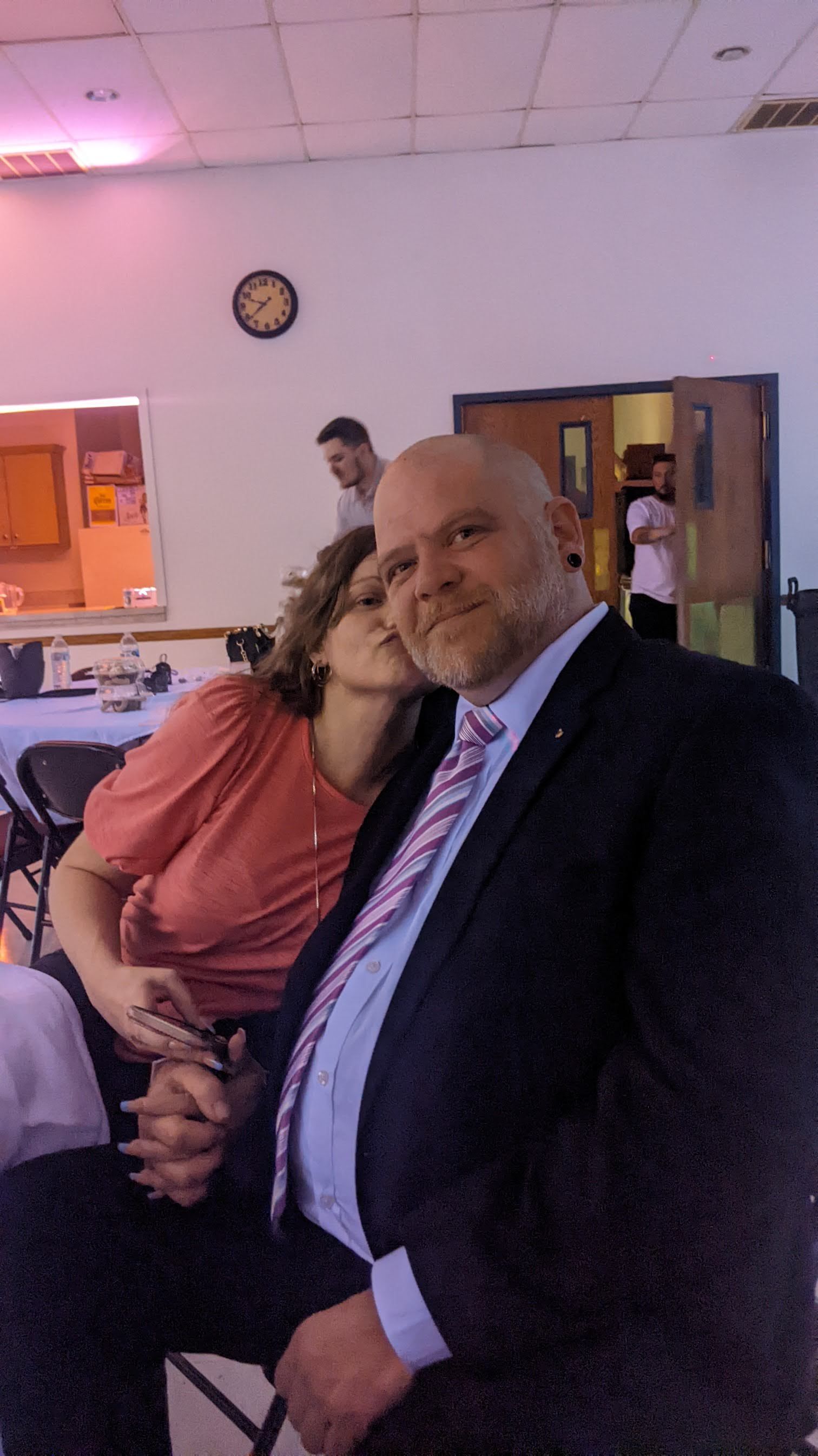 Two people posing closely at an indoor event, wearing formal attire, with tables and other guests in the background.