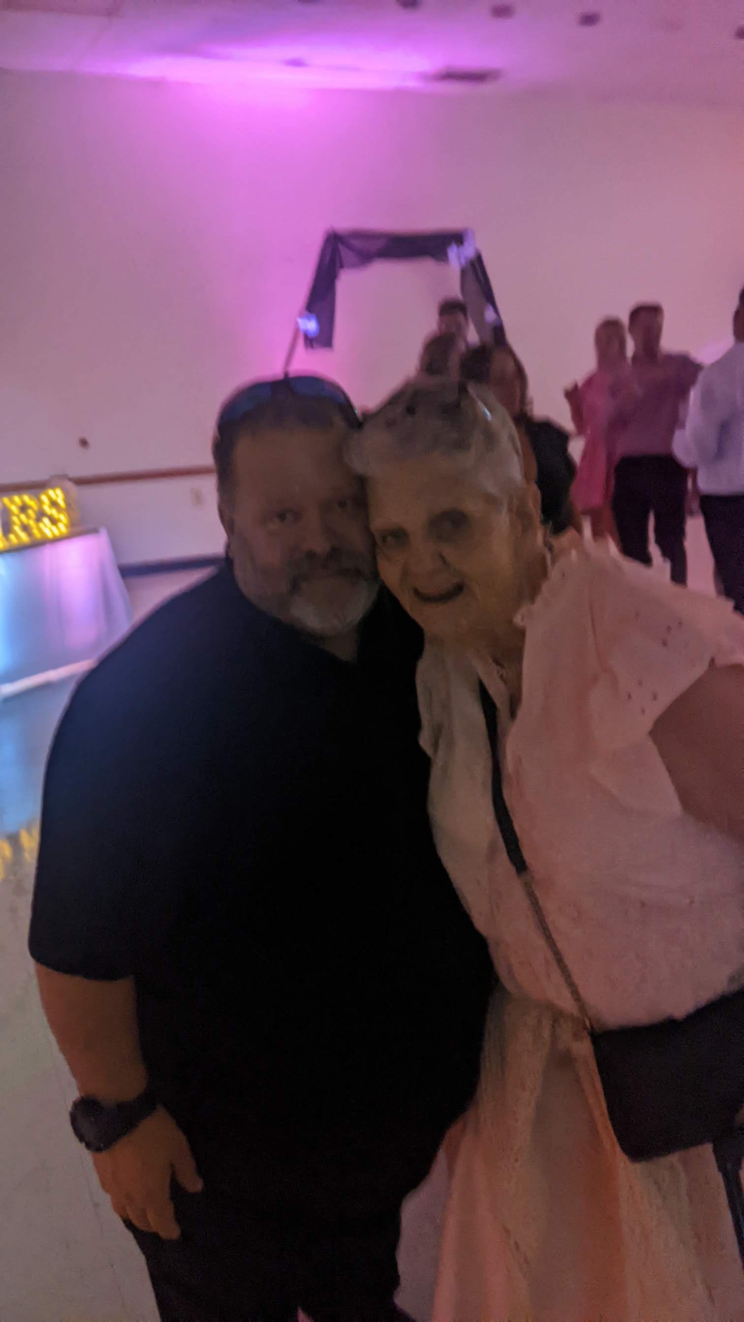 Two people posing together at a dimly lit event, one in a pink dress and one in a black shirt.
