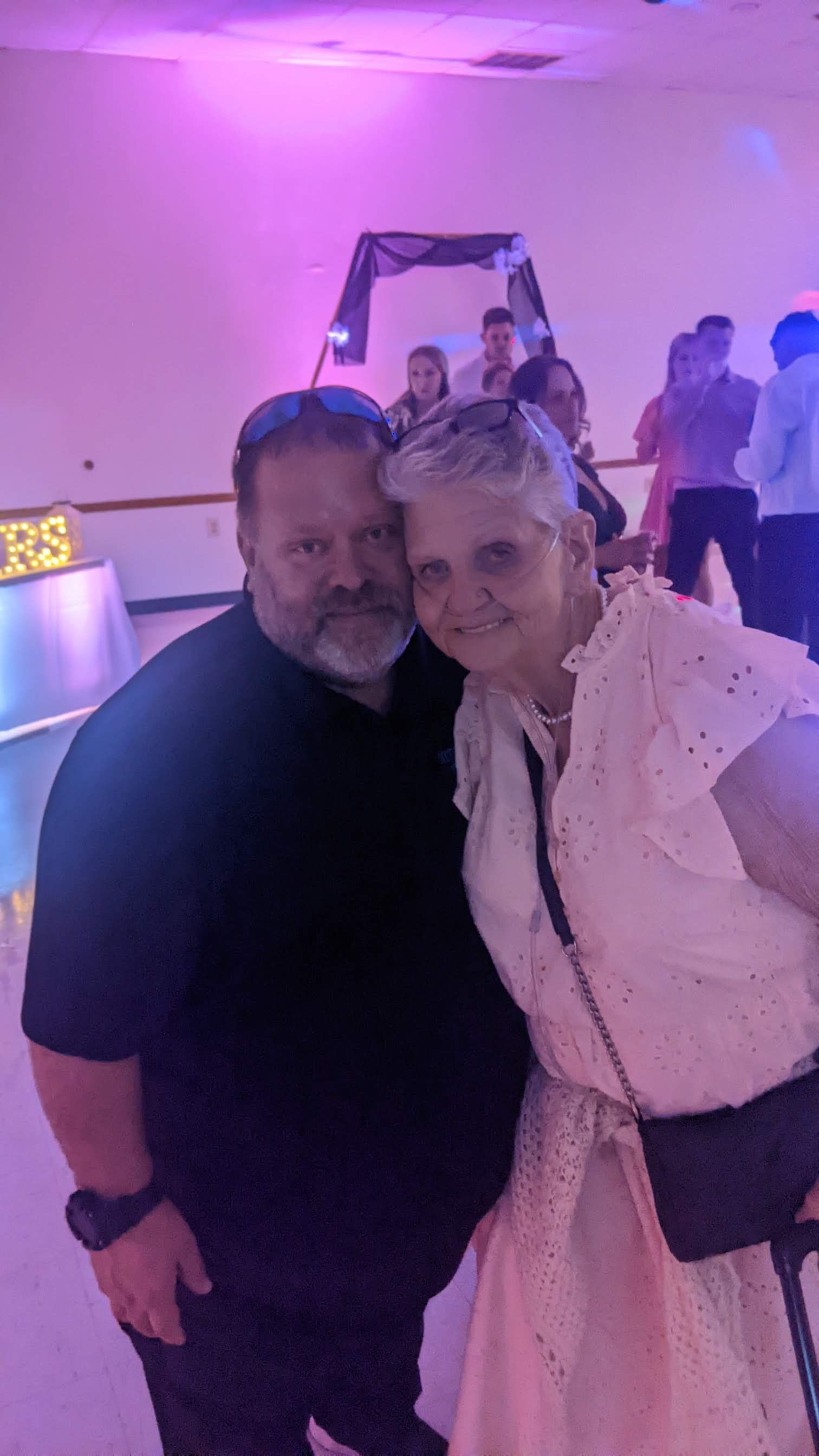 Two people posing closely in a purple-lit party room, one in a black shirt and one in a light dress.