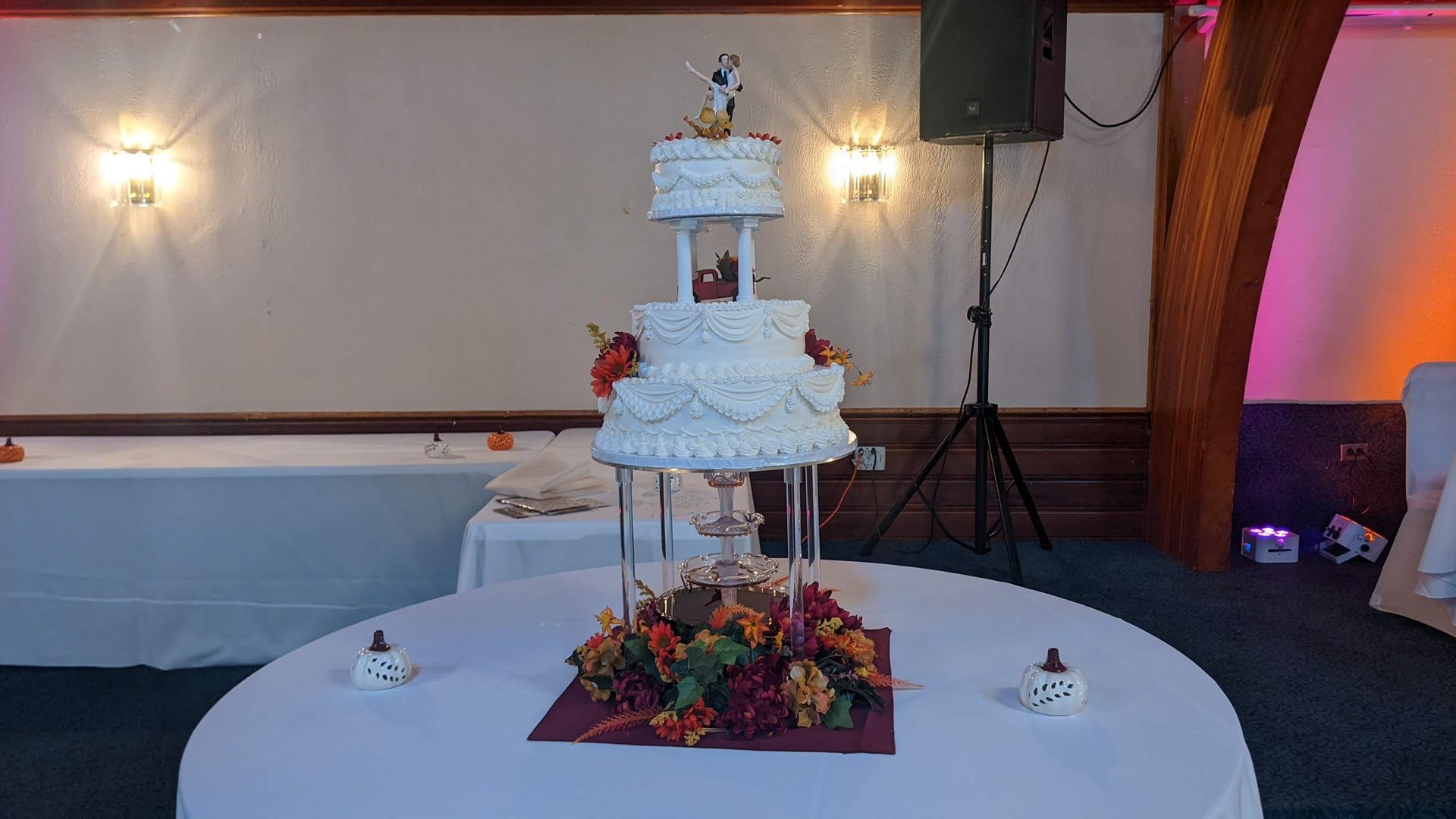 White tiered wedding cake on a decorated table in a reception hall with floral centerpiece and wall lights