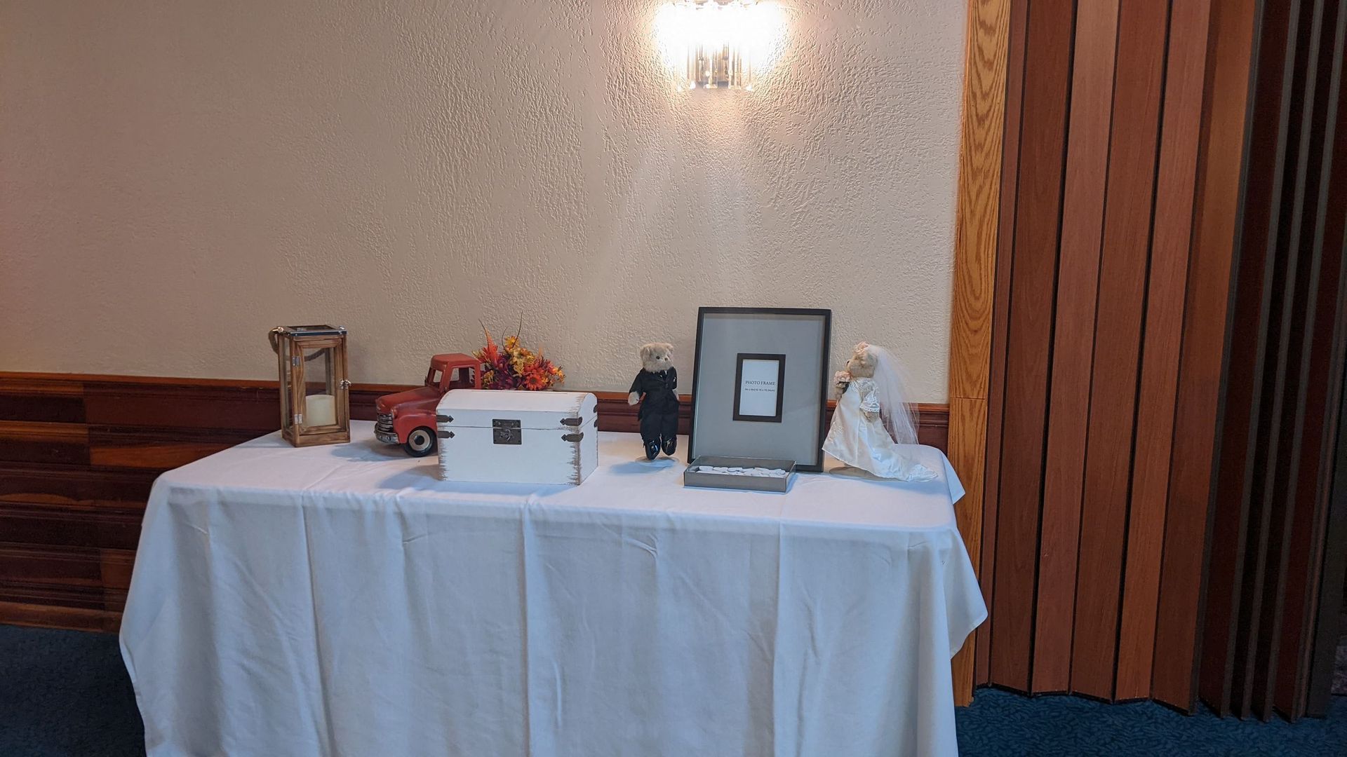 Table with framed photos, a lantern, white box, flowers, and a doll against a beige wall in a room.