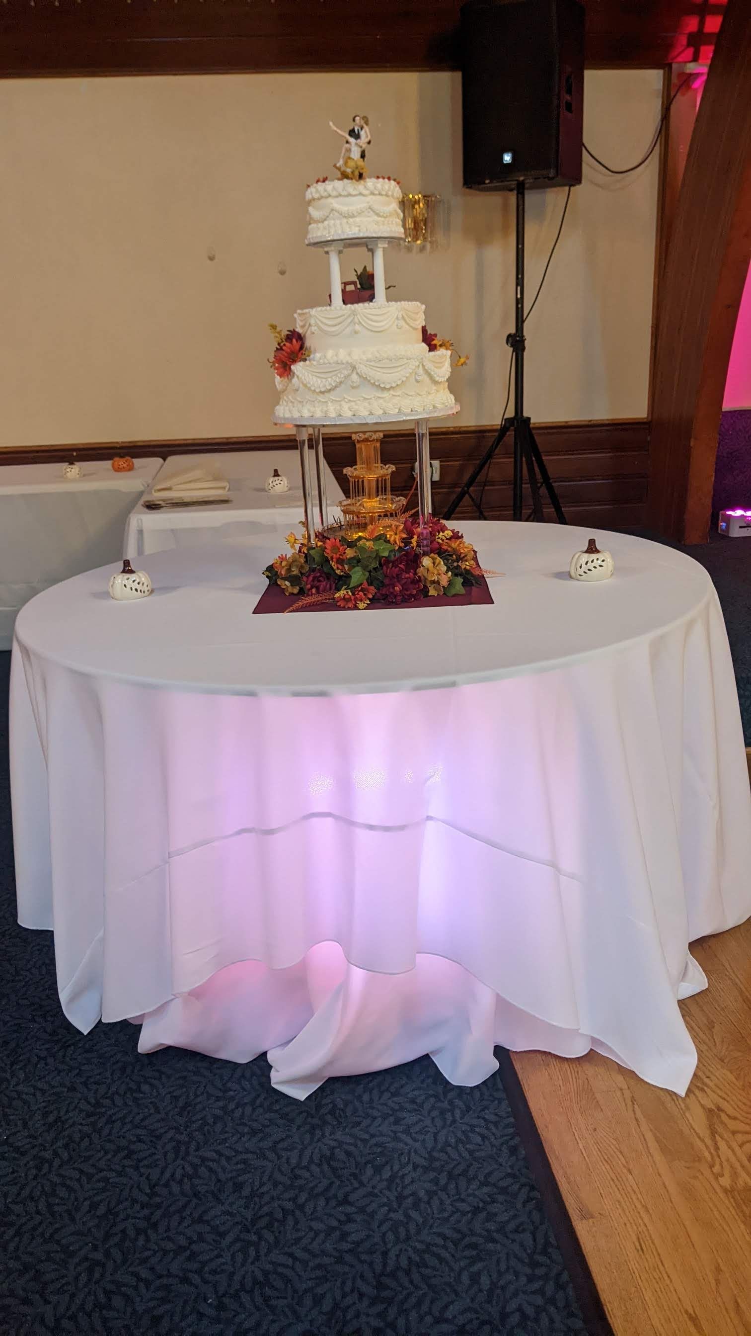 Wedding cake on a white-draped table with floral centerpiece and soft pink uplighting in a reception hall