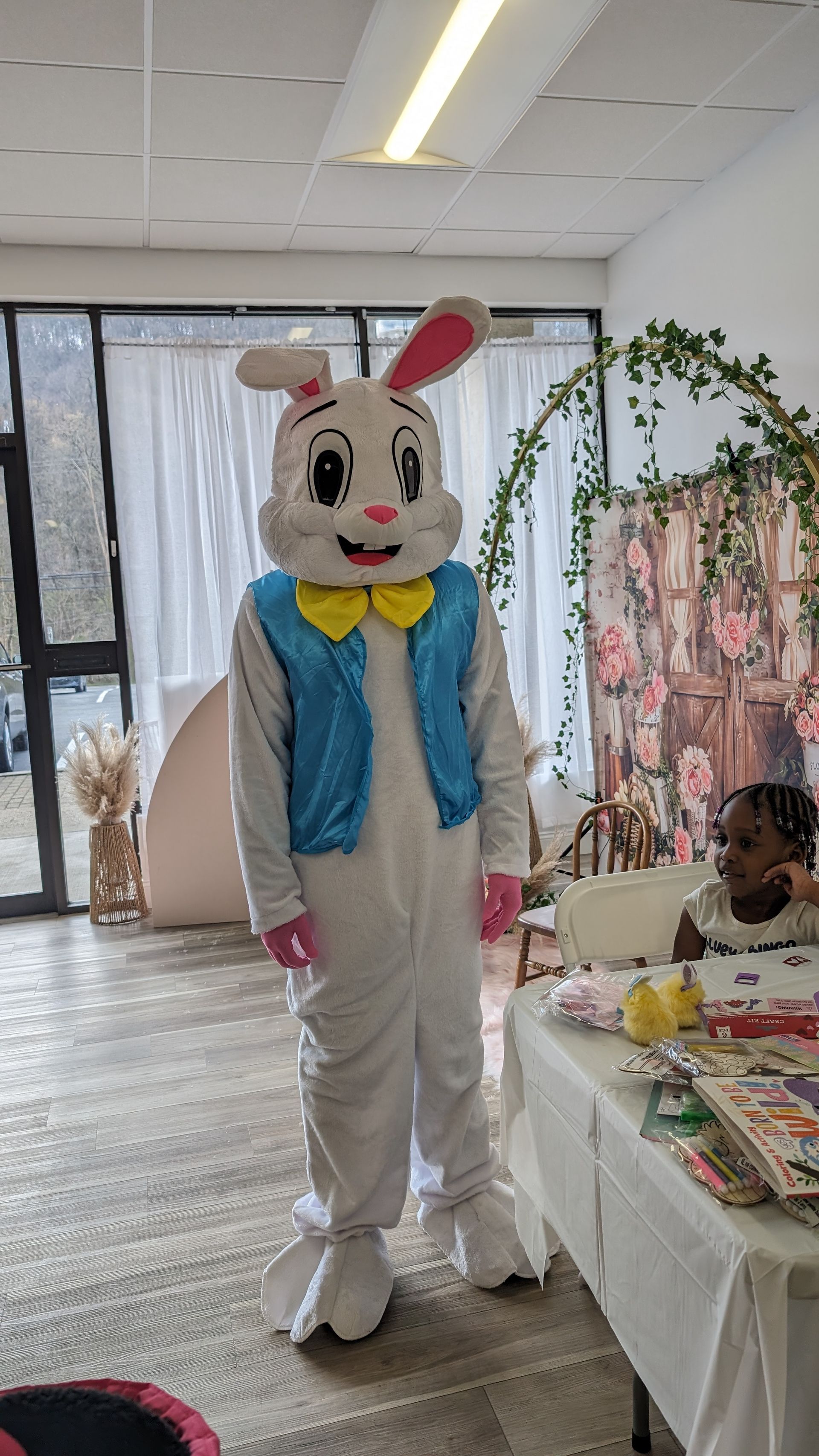 Person in a white bunny costume with pink ears and blue scarf stands in a bright room beside a table.