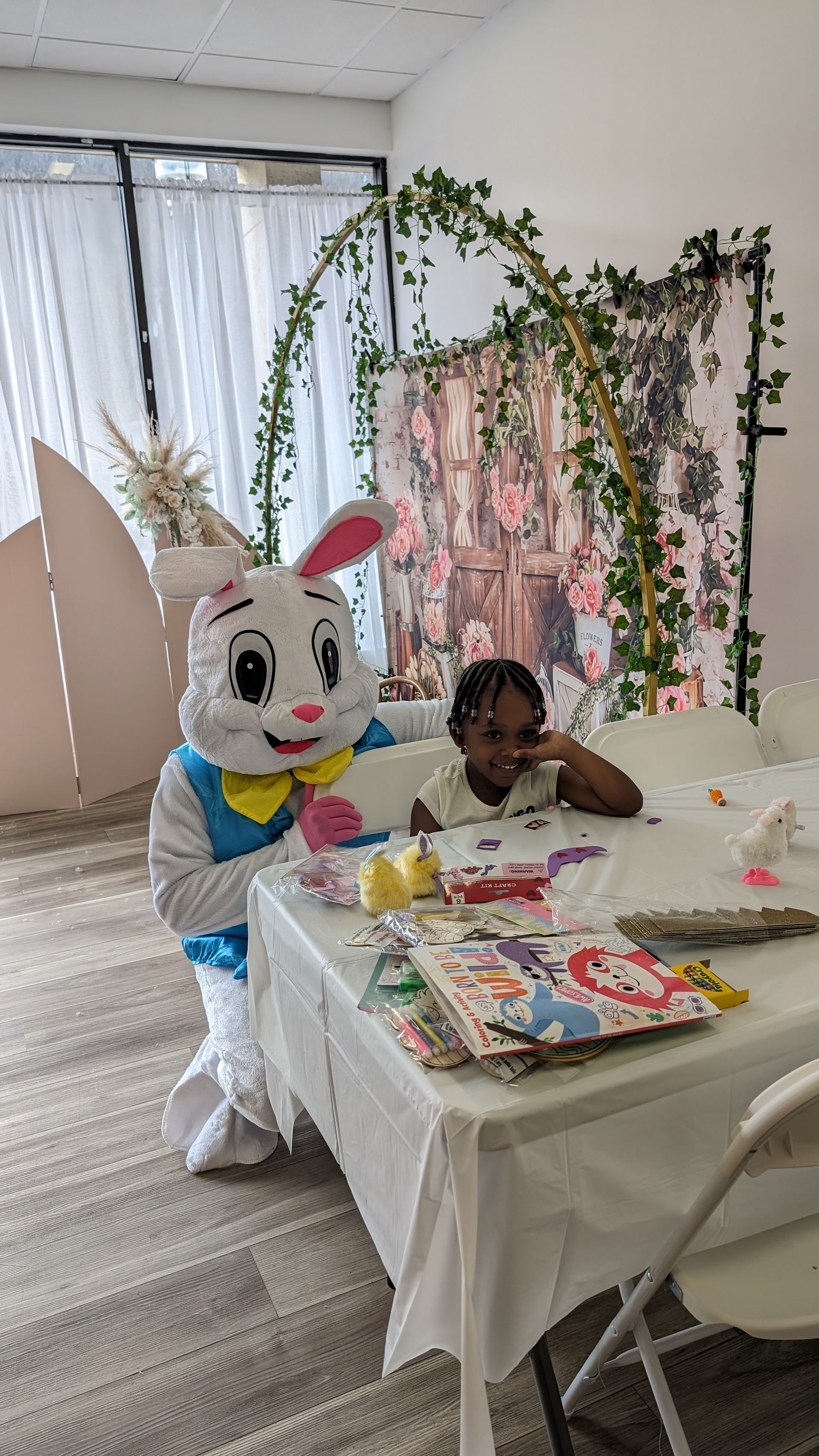 Child with a large white bunny mascot at a decorated table in a bright room.
