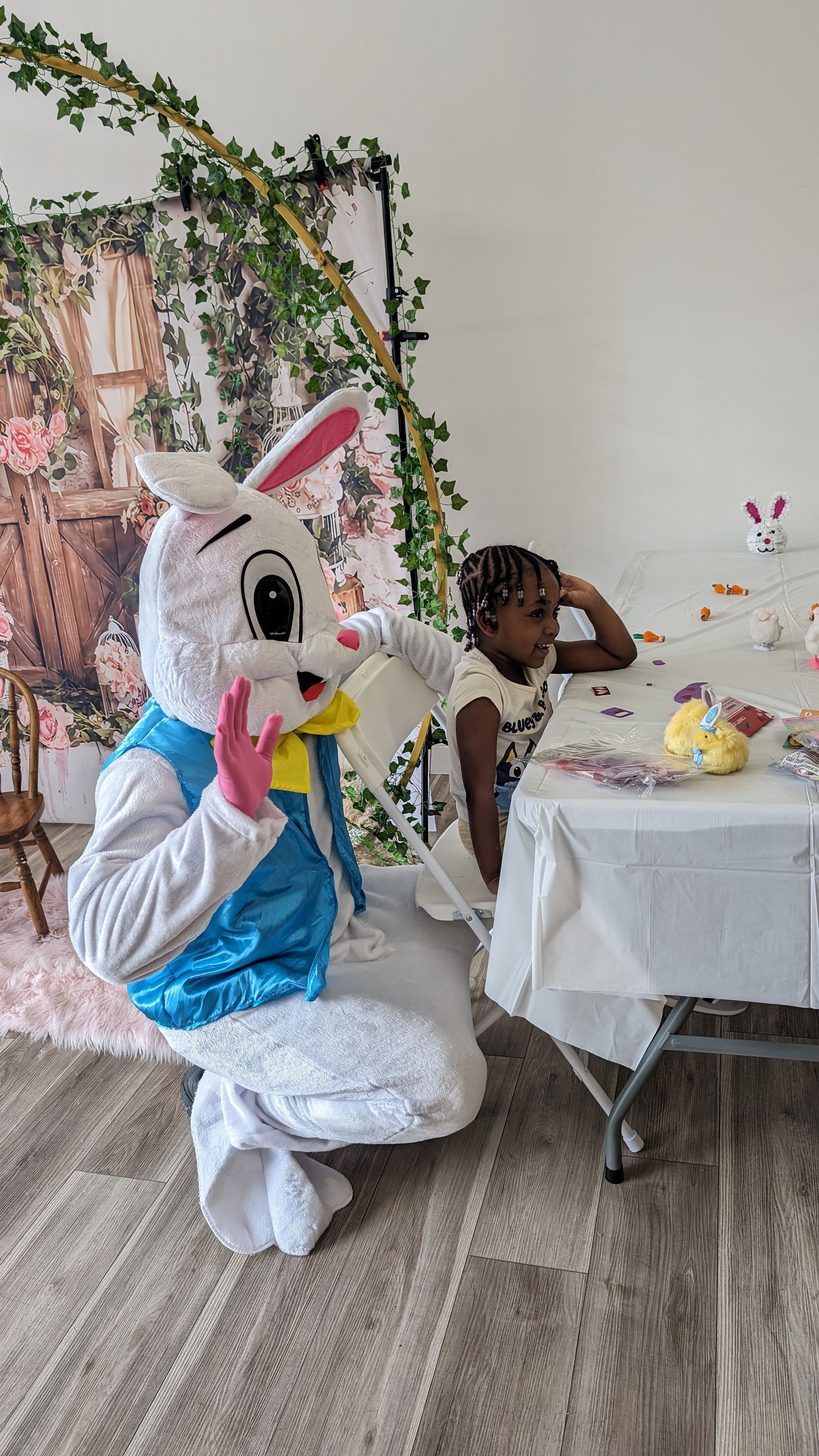 Person in a bunny costume kneels beside a child at a decorated party table indoors.