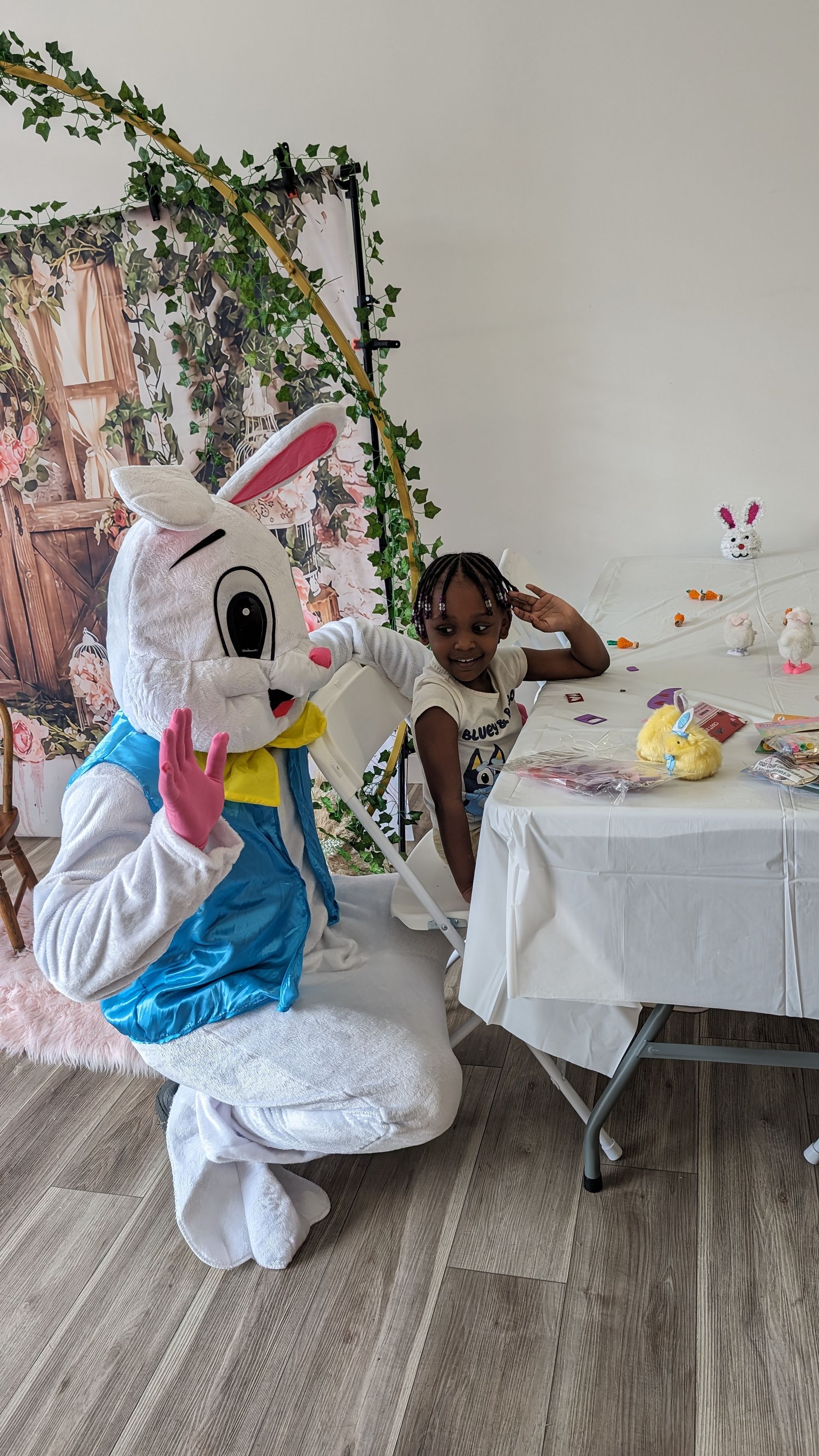 Person in a large bunny costume at a decorated table in a party room, with a child posing nearby.