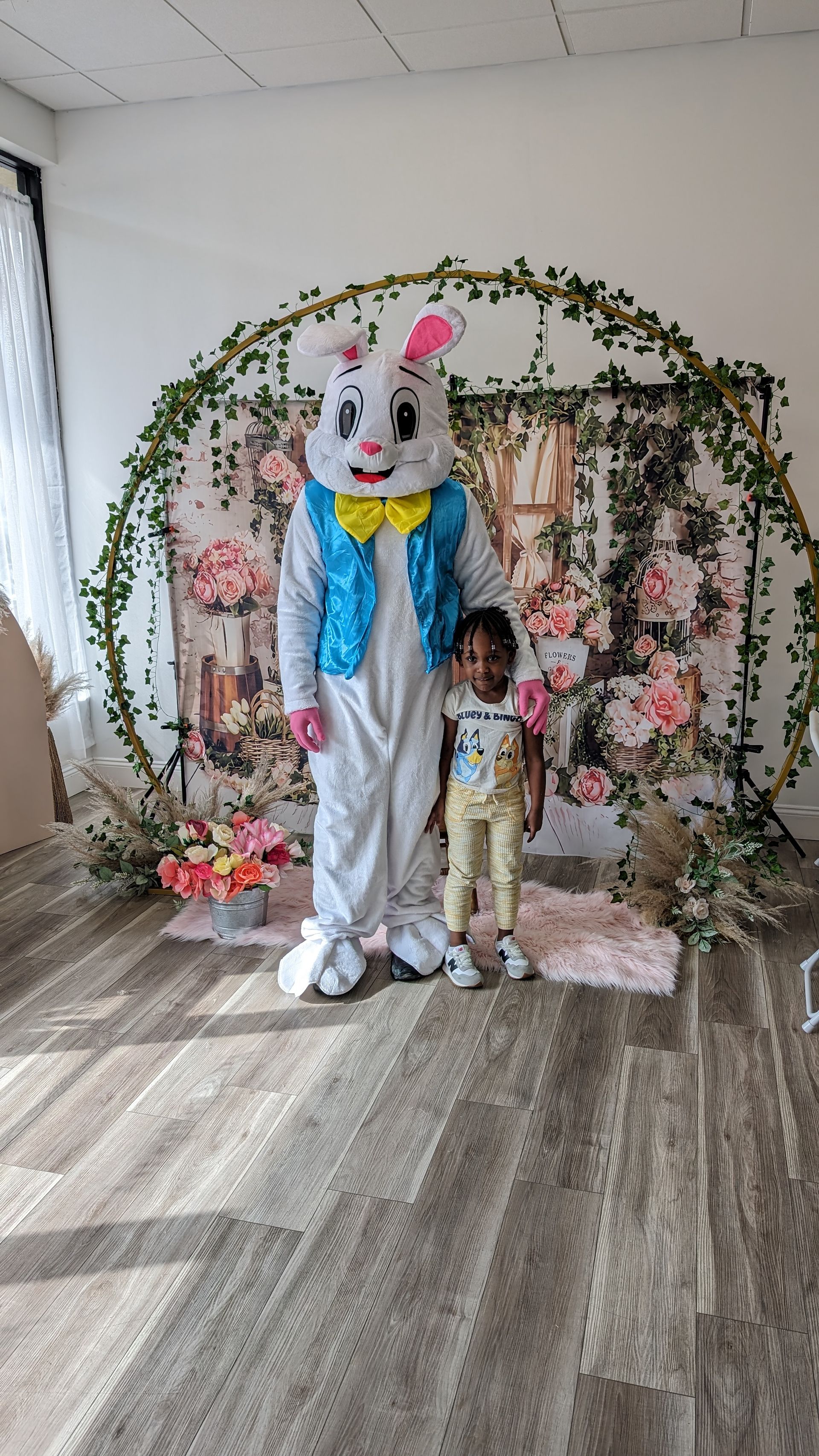 Child standing beside a person in a white bunny costume under a floral arch indoors