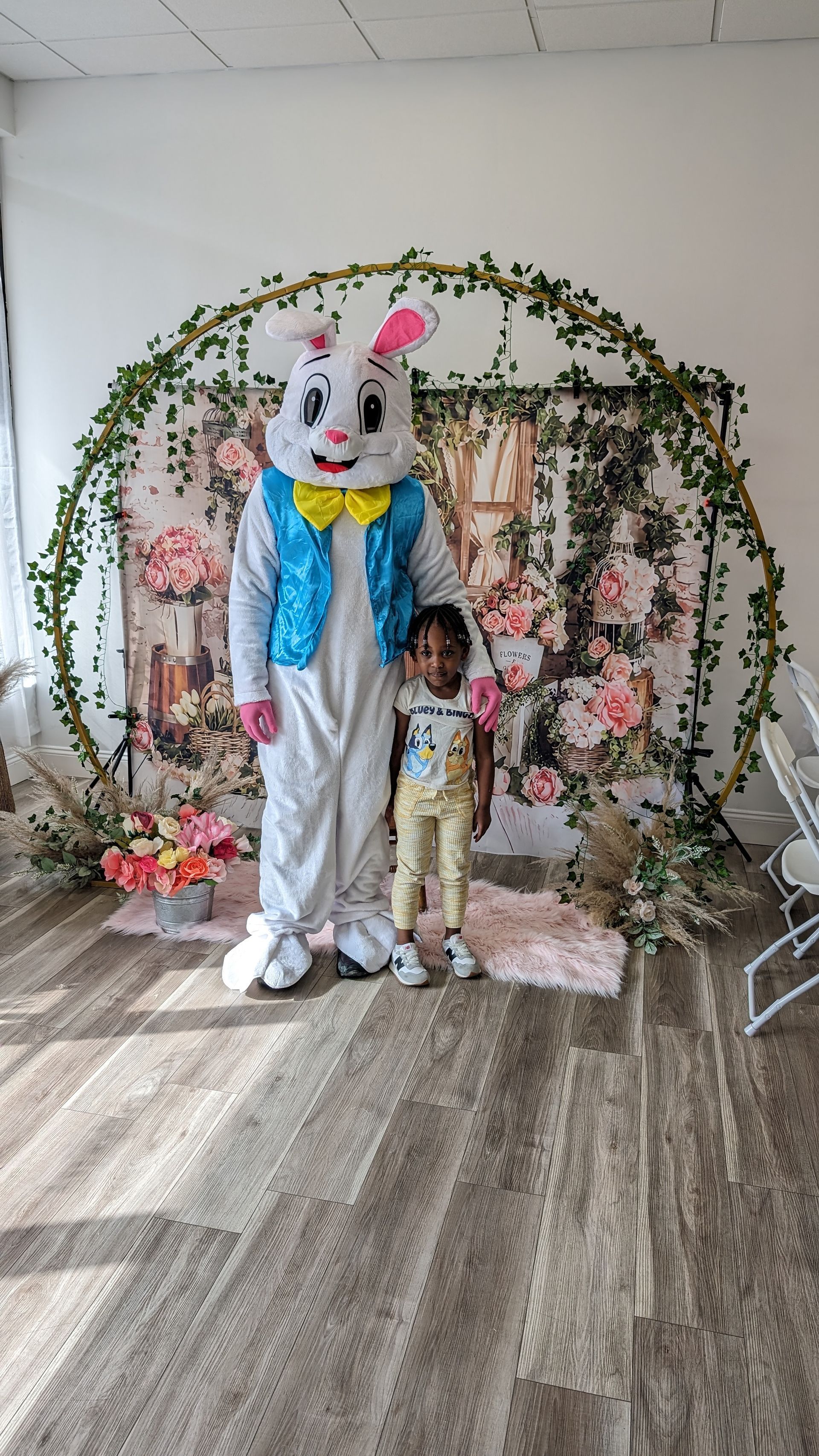 Child standing beside a person in a white bunny costume under a floral arch with pink decorations