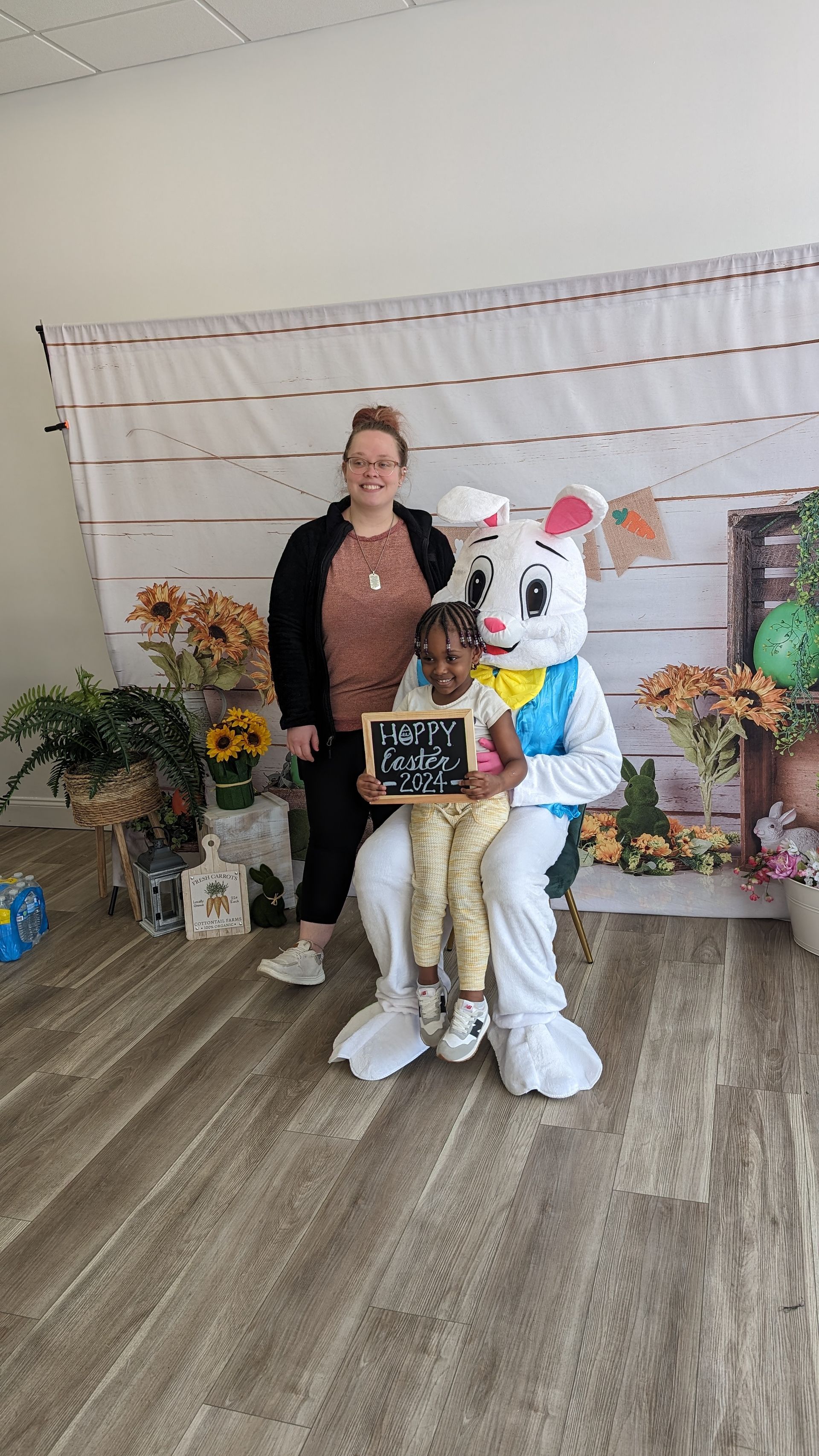 Woman with a child in a white mascot costume holding a chalkboard, standing in a decorated indoor photo area.
