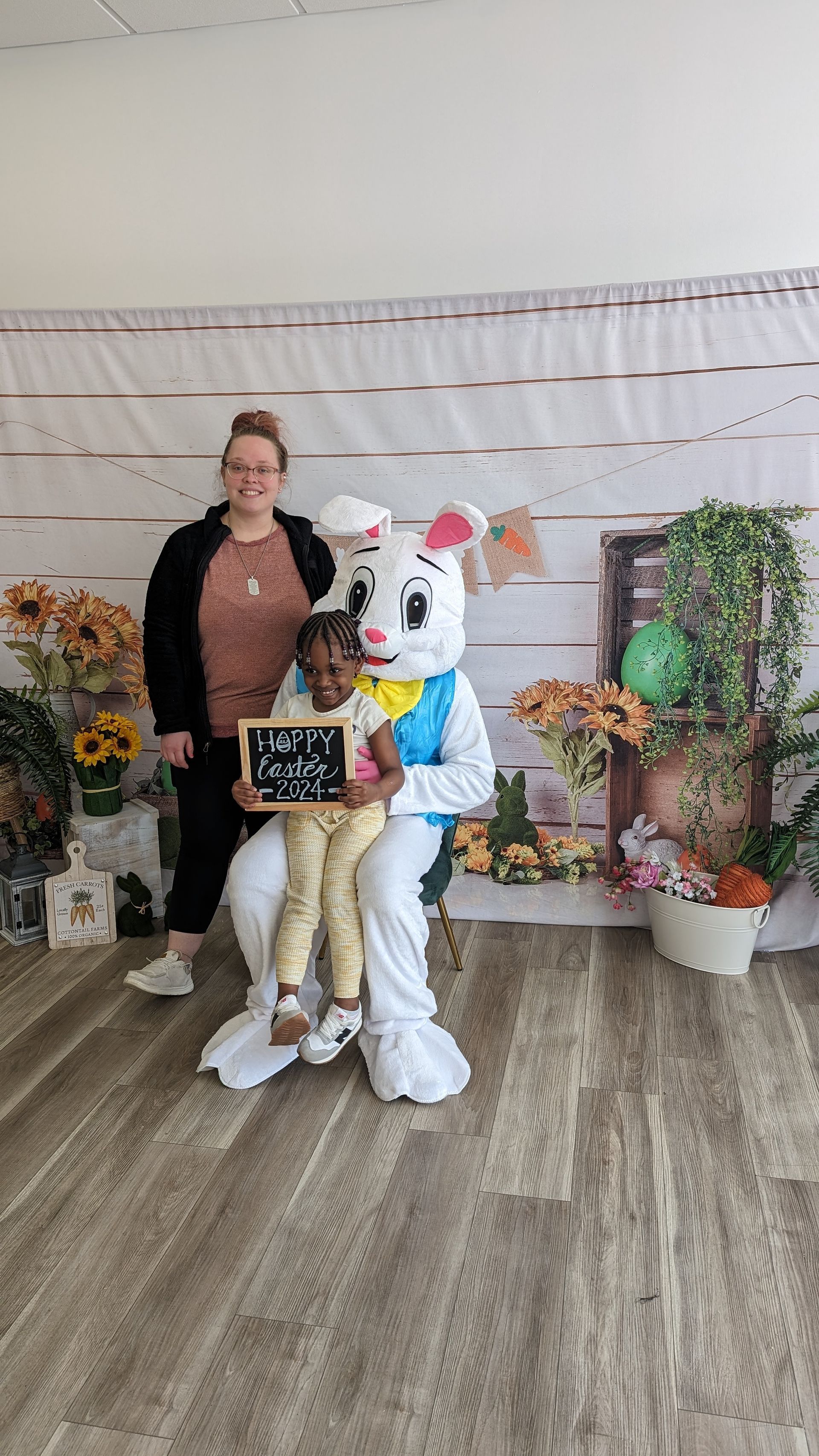Woman and child holding a sign beside a colorful bunny mascot in a decorated indoor photo area