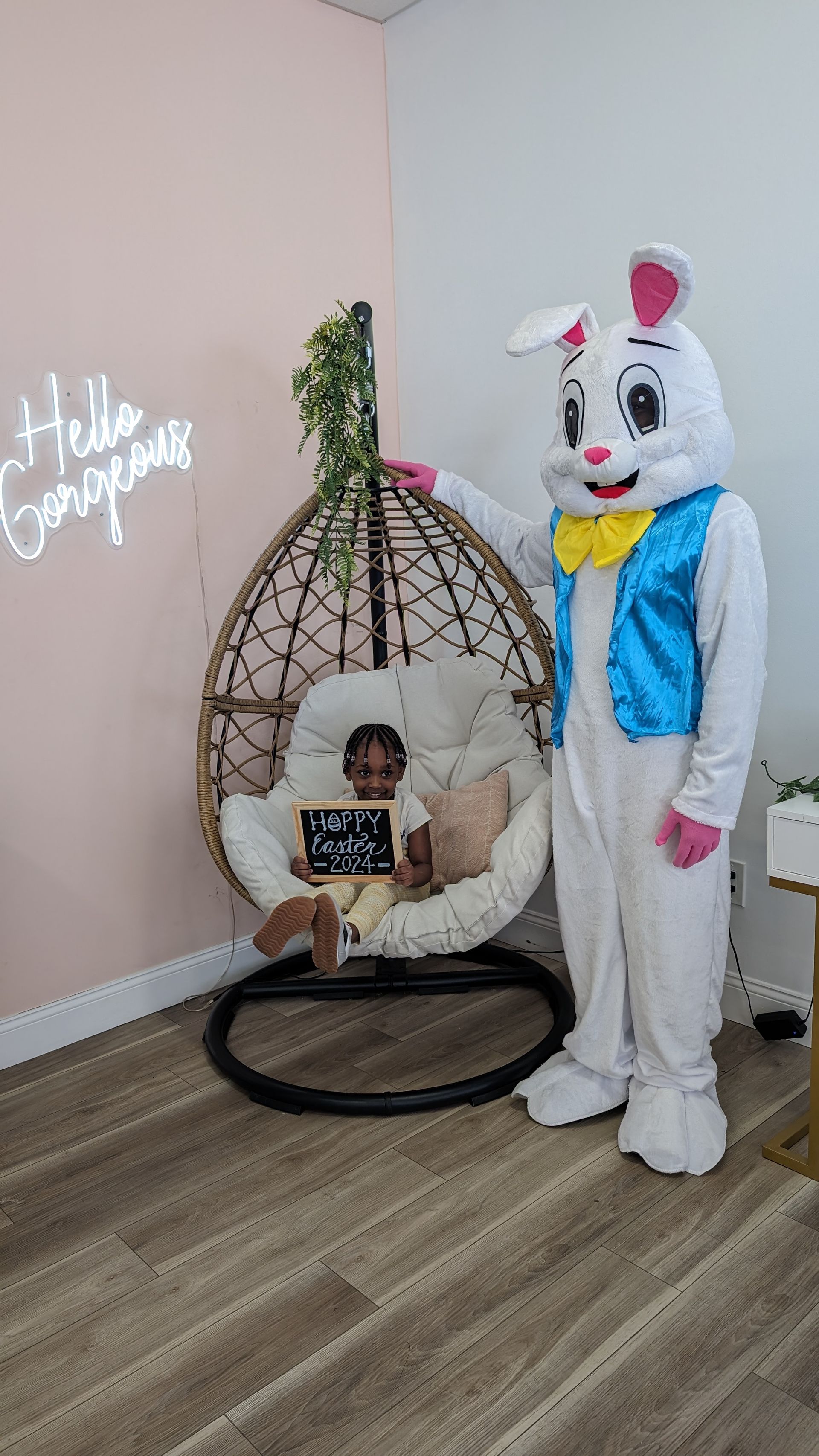 Person in white bunny costume stands beside a baby in a hanging wicker chair, with “Hello Spring” wall decor.