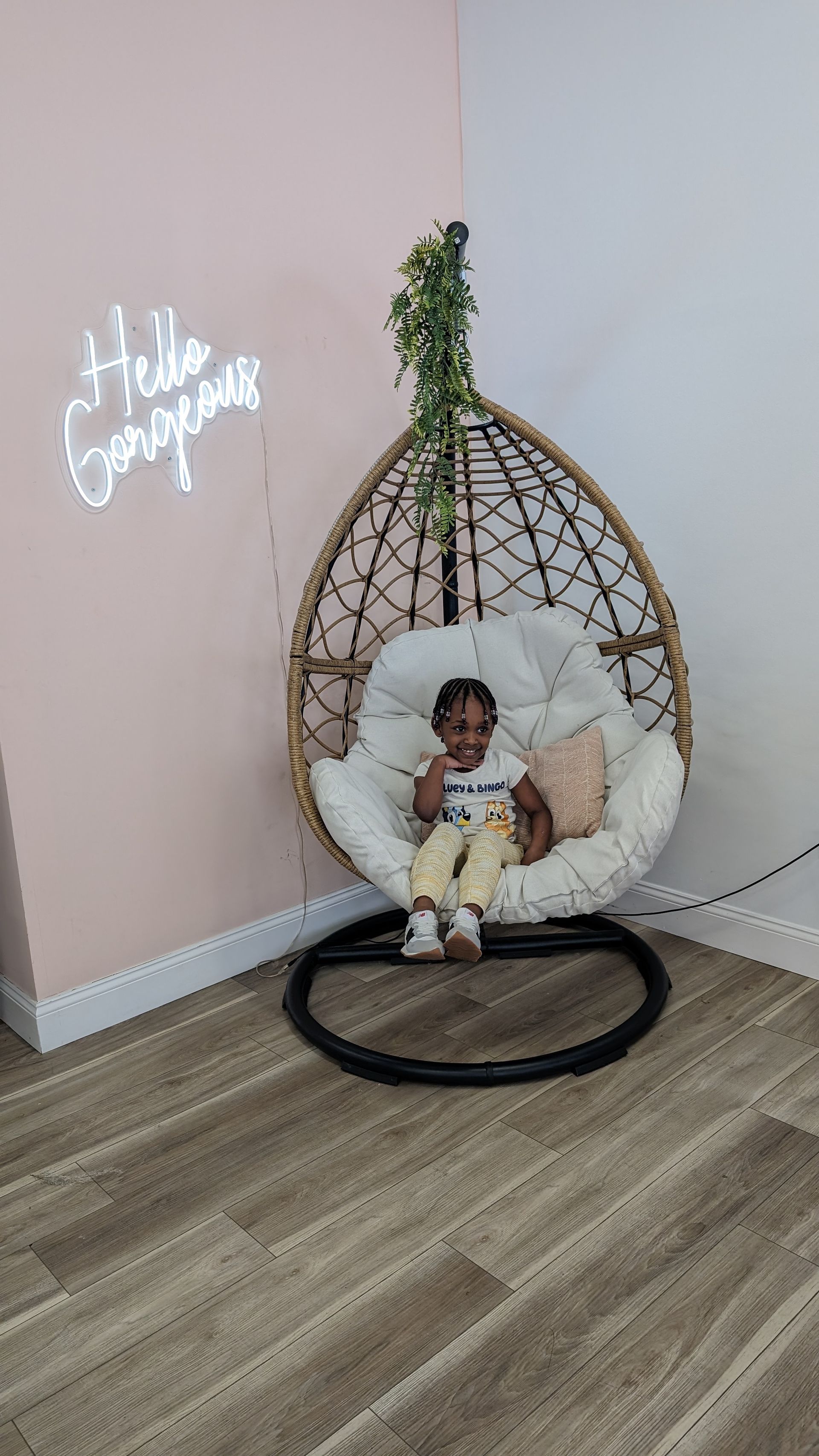 Child sitting in a wicker hanging chair with pillows in a pastel room, beside a glowing wall sign.