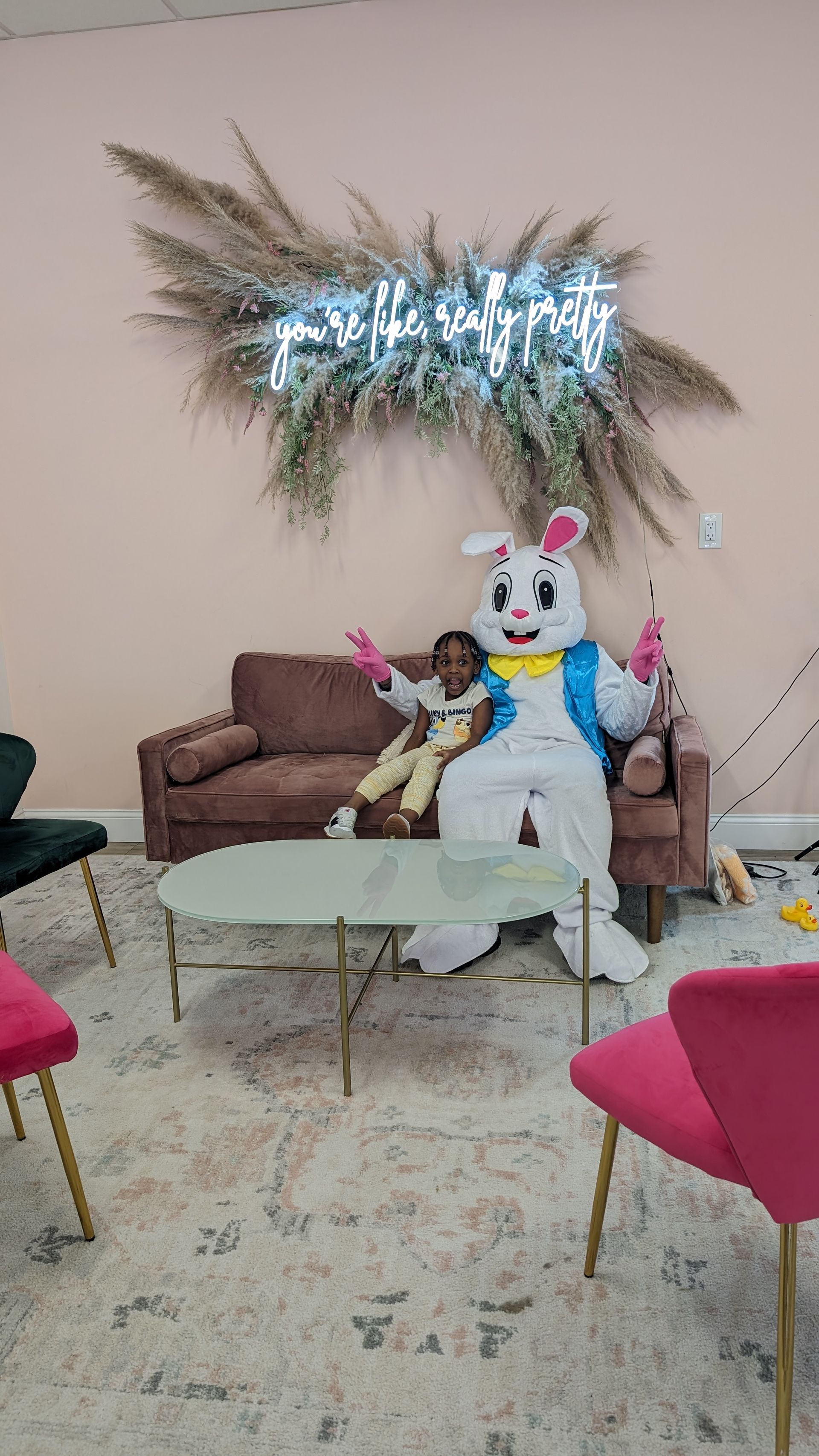 Child on couch beside large rabbit mascot in decorated living room with balloons and metallic wall decor