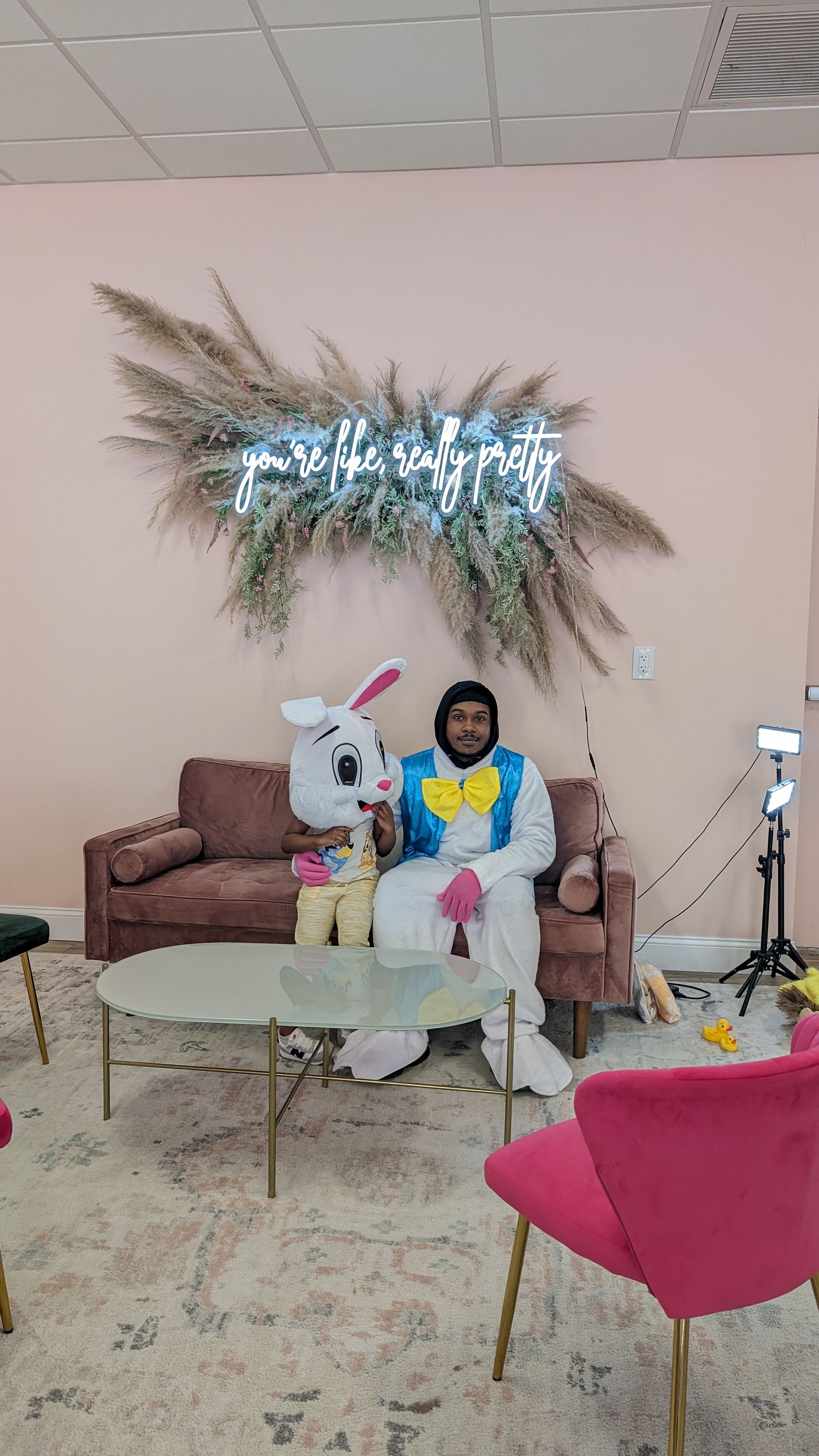 Person sitting on a brown sofa in a decorated living room with a glass table and pink chair.