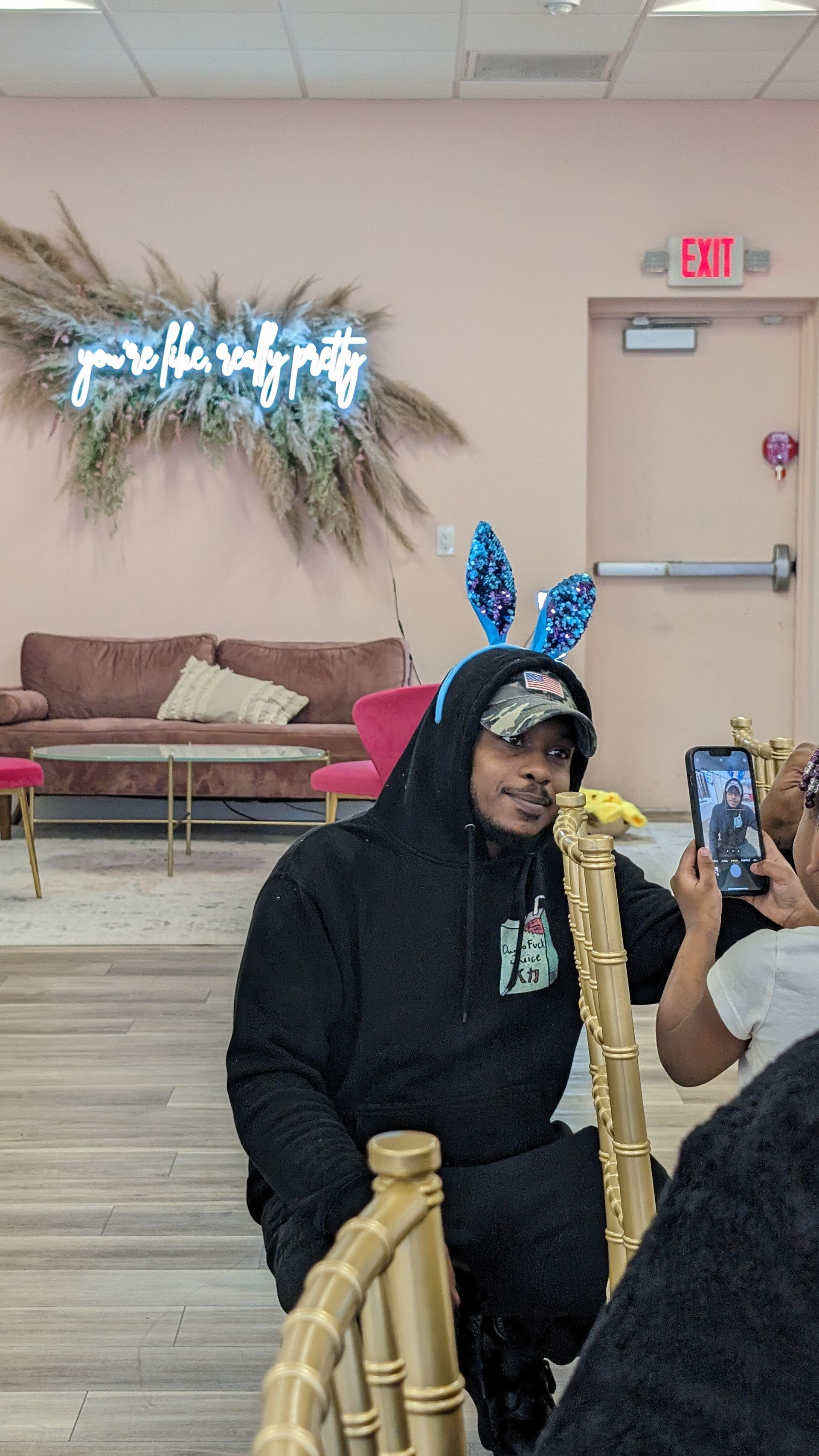 Person in black hoodie with blue bunny ears smiles for a photo in a lobby decorated with balloons.