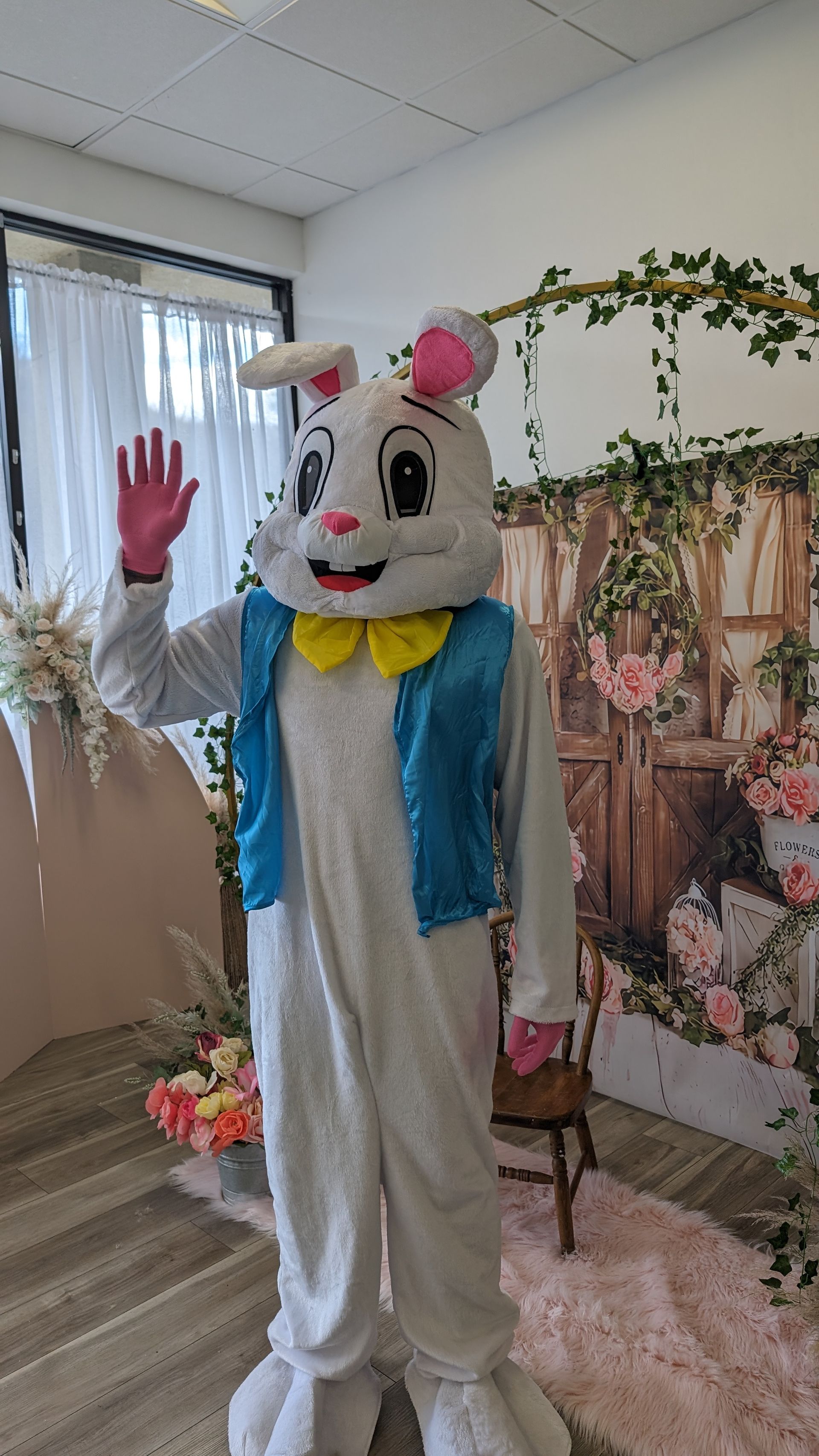 Person in a white bunny costume with pink ears and blue vest waving indoors at a decorated event backdrop
