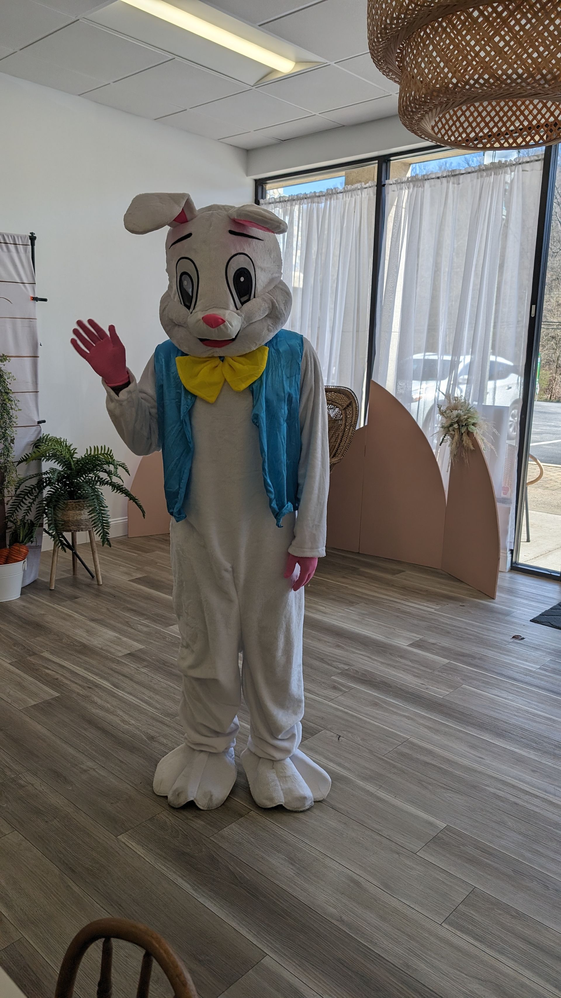 Person in a white bunny costume with a blue scarf and yellow bow waving indoors.