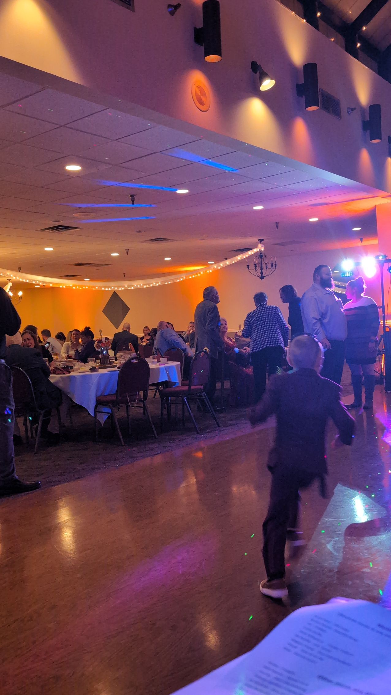 Dimly lit banquet hall with seated guests, warm orange lighting, and a dance floor with people mingling.