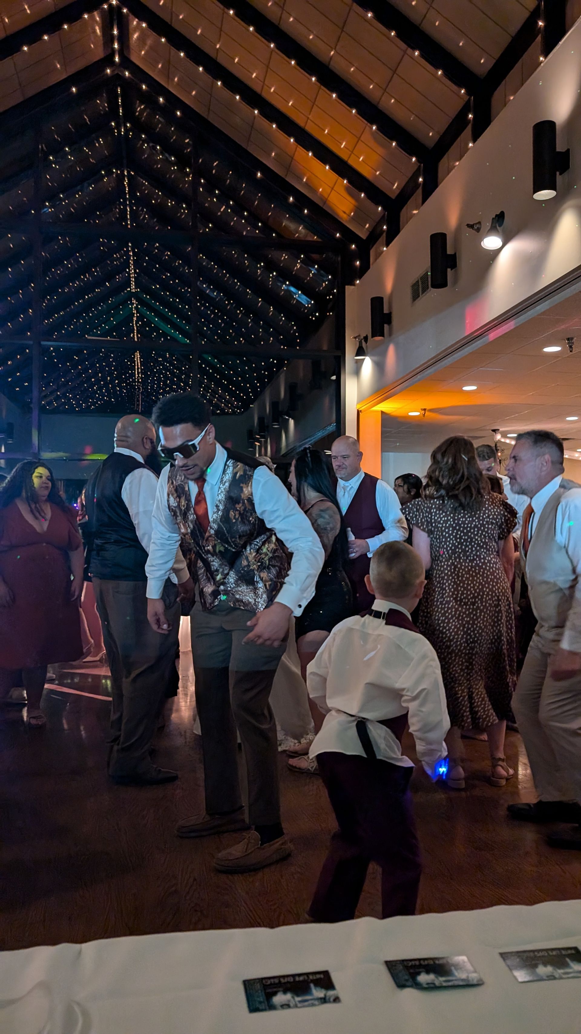 People mingling and dancing in a warmly lit indoor hall with a high wooden ceiling.