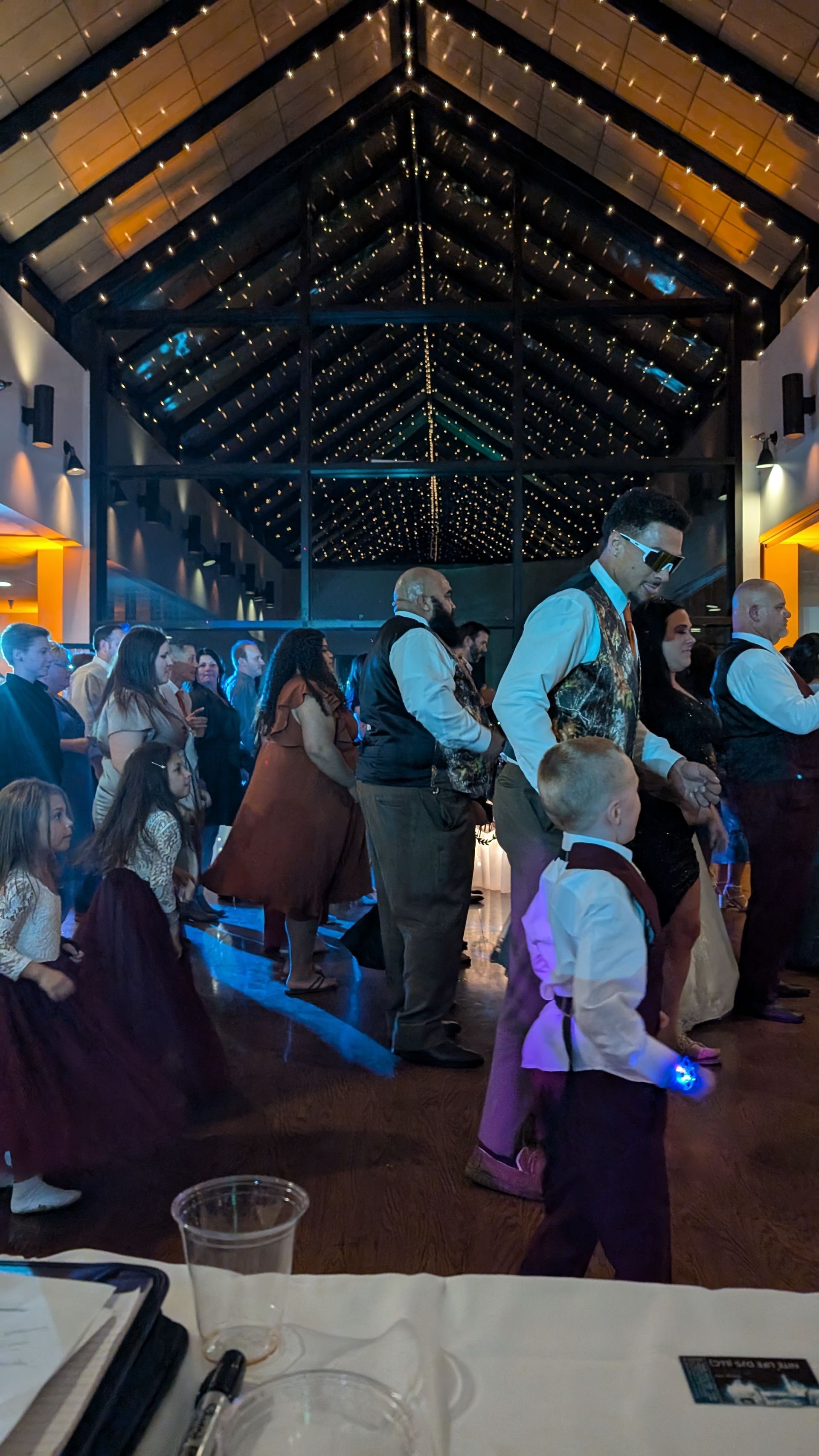 People standing and dancing in a dimly lit banquet hall with string lights and blue-purple ambient lighting
