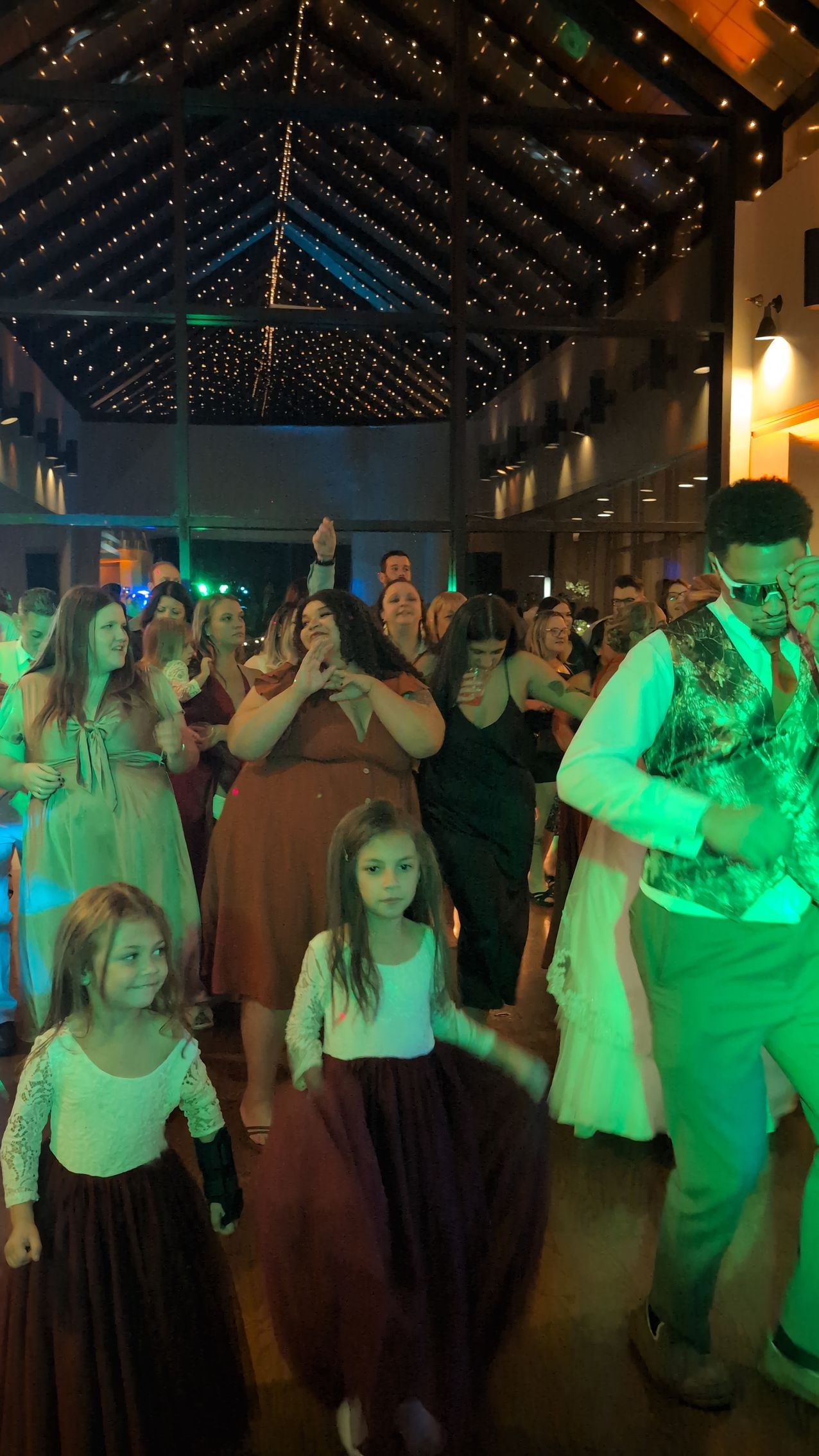 People in formal wear dancing at a warmly lit indoor party or wedding reception.