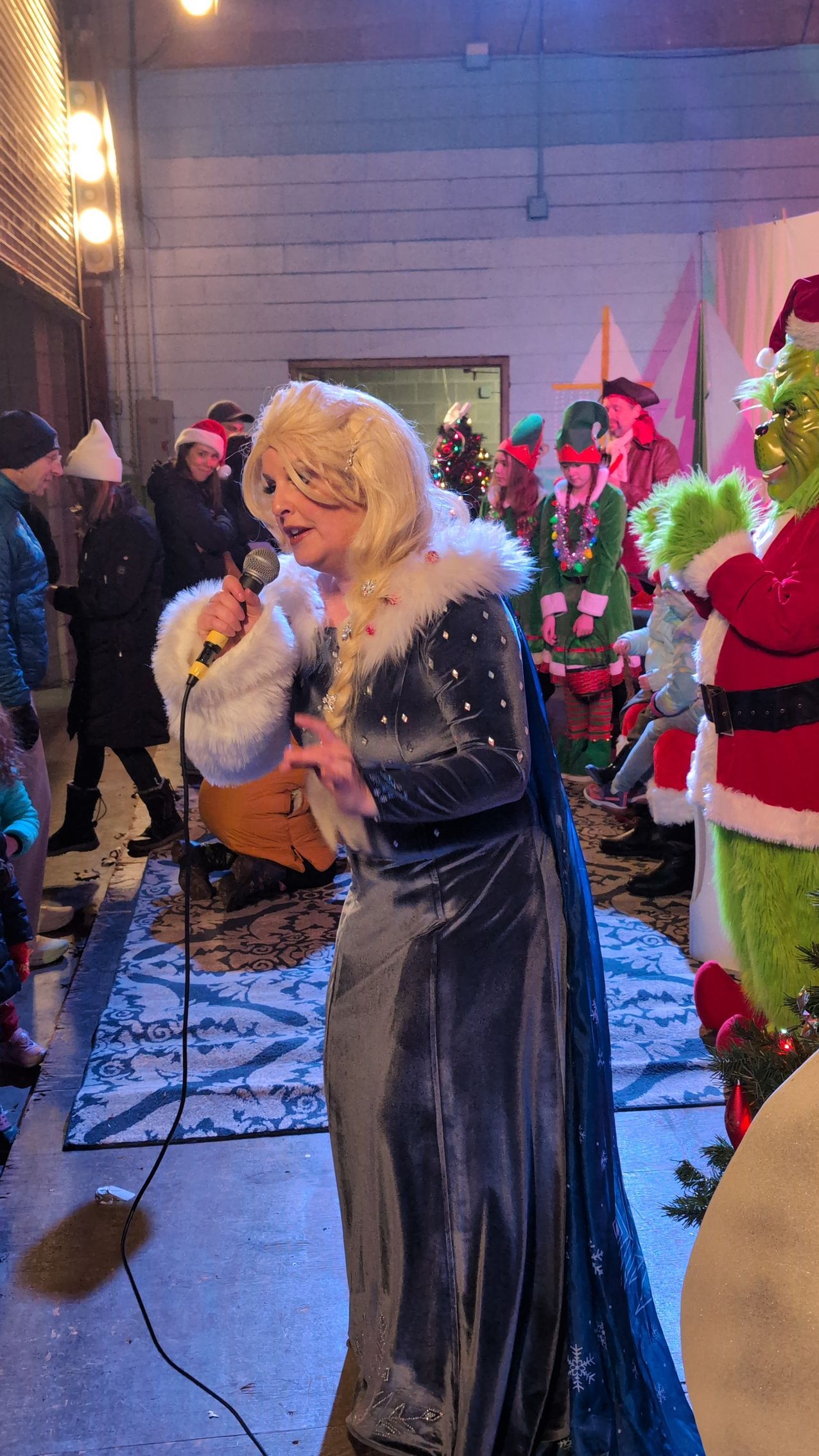 Woman in a long silver gown singing into a microphone at a festive indoor event, with decorations and Santa nearby