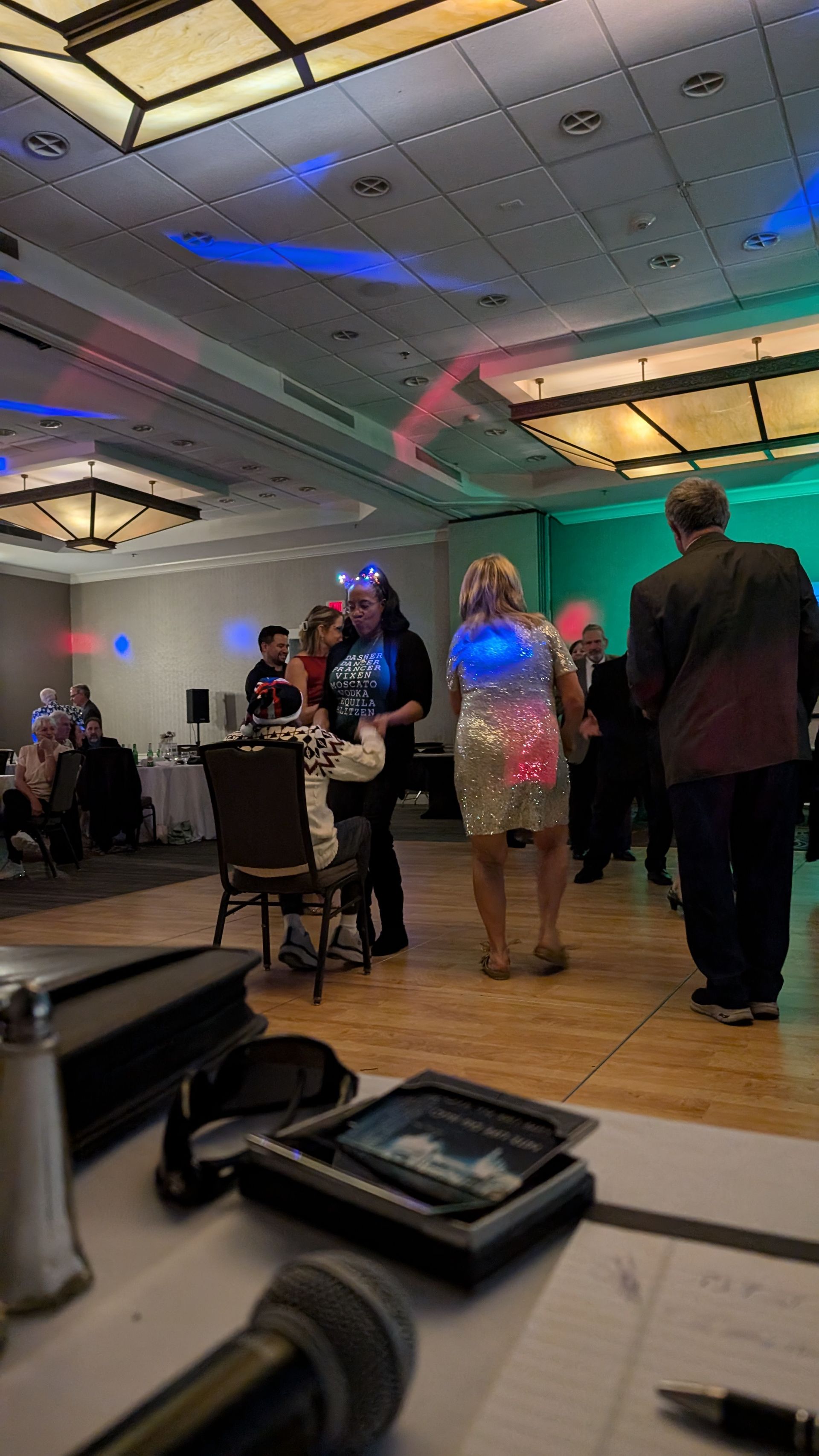 People standing and talking in a banquet hall with colorful ceiling lights and tables.